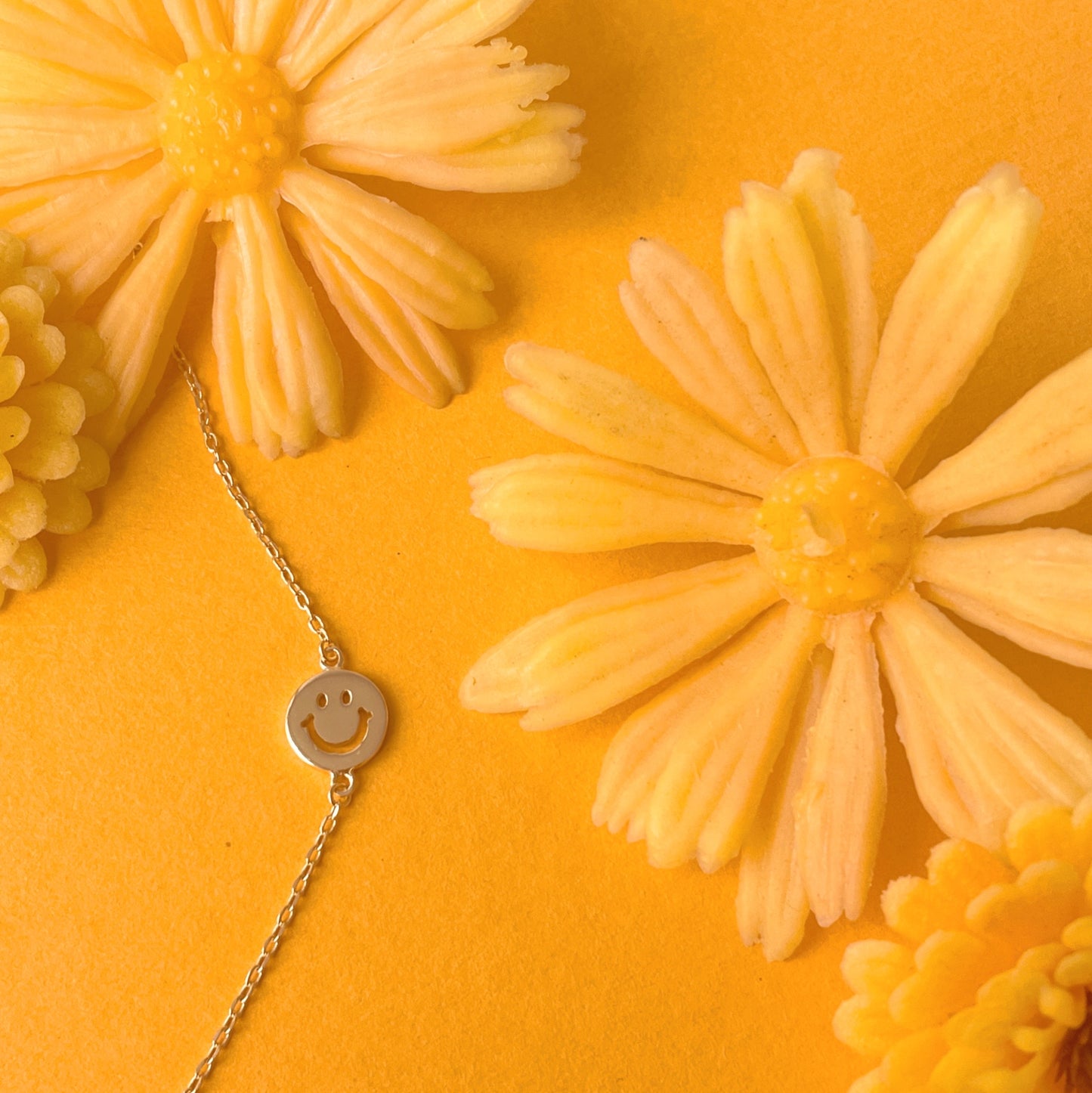 Gold chain necklace complete with classic smiley face pendant turned sideways. The pendant is connected to the chain by two loops on each side. This image features the necklace resting on a marigold colored ground with fake yellow daisies surrounding it.