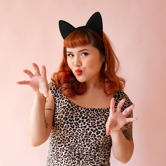 Model in leopard body suit with red hair wears a black glitter cat ears headband.