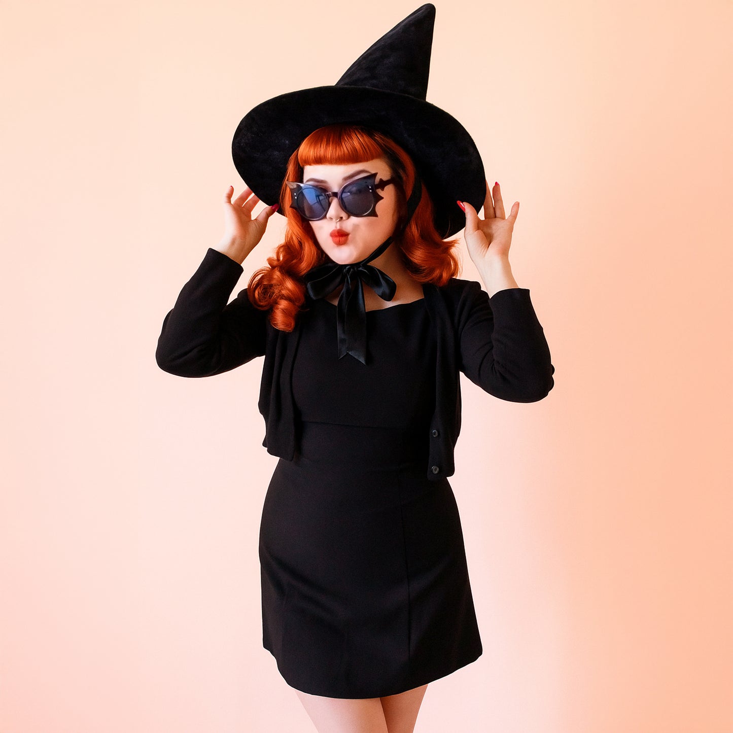 A red headed woman wears black bat shaped sunglasses and a tall black witch hat with a bow at the neck. She is wearing an all black outfit.