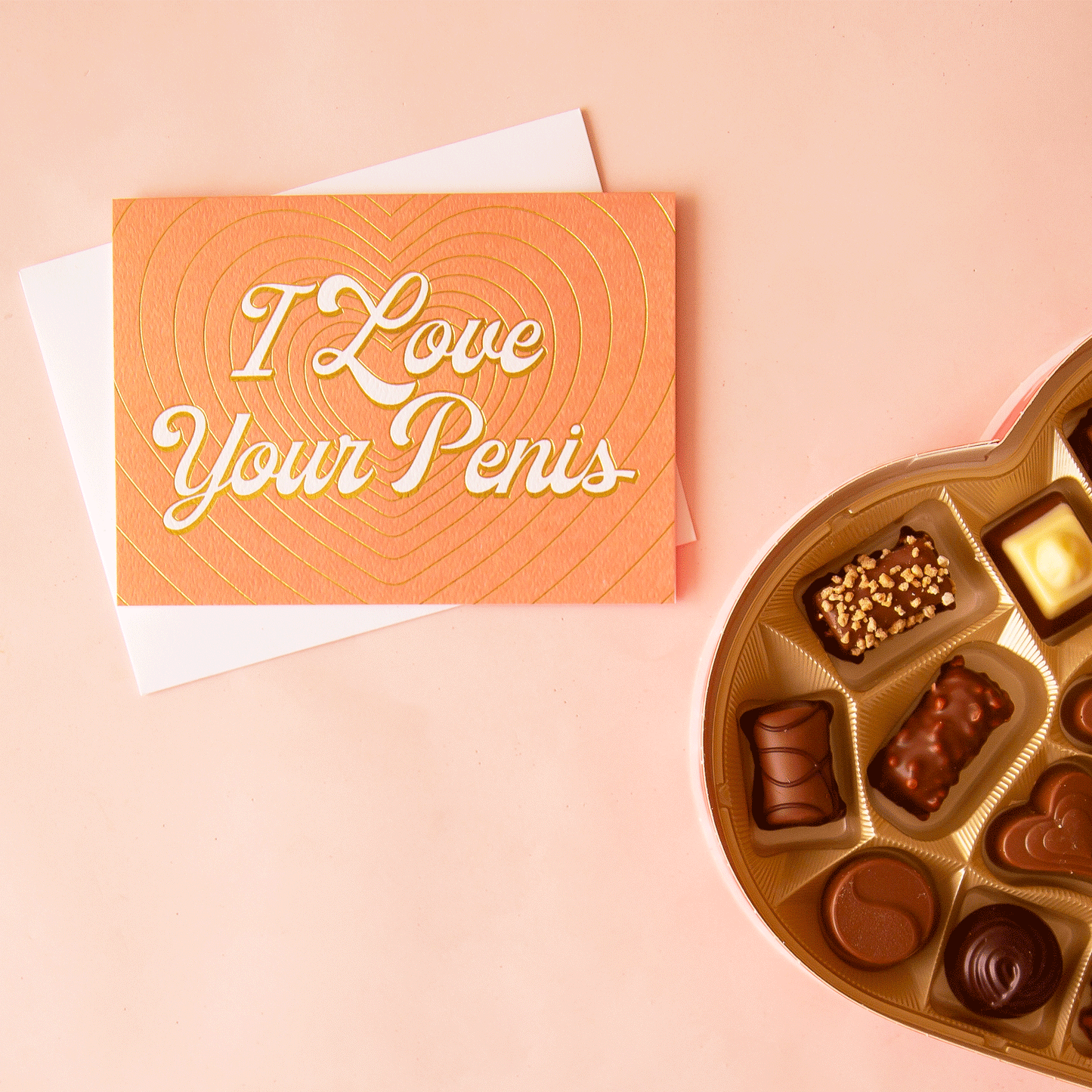 On a pink background is a peachy colored card with gold foiled layered heart outlines with ivory text in the center that reads, "I Love Your Penis".