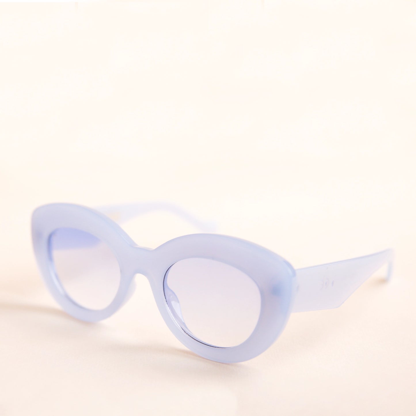 On a neutral background is the Gemma Sunglasses in a light blue shade. They have a rounded shape with a slight cateye flair at the corners. The frame material is a durable plastic and the lenses are a similar tone.