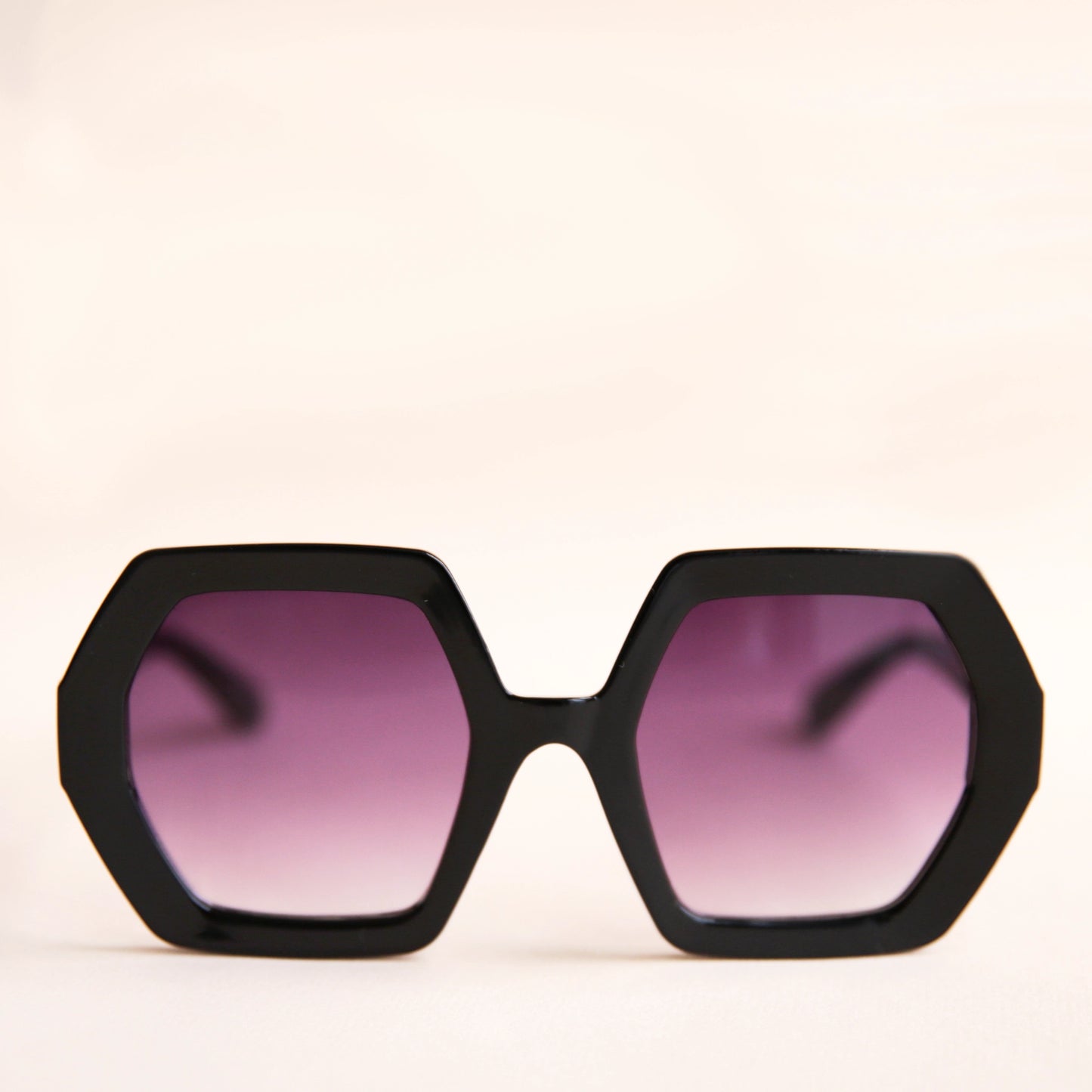On a neutral background is a pair of hexagon shaped black sunglasses with purple lenses.