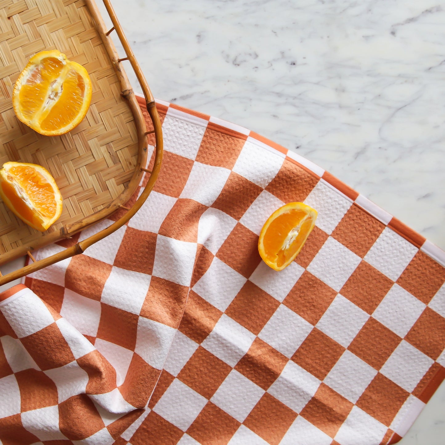 On a marble backdrop is an orange and ivory checkered kitchen towel with a staged orange cut up next to it.