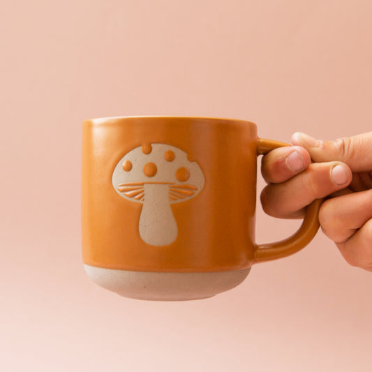 On a pink background is a ceramic mug with a light colored mushroom design.