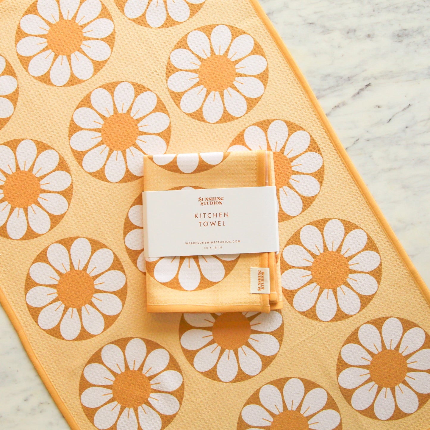 On a marble background is a yellow and white daisy print kitchen towel with a subtle waffle texture along with the folded towel in the center to show how the item arrives. One towel per purchase.