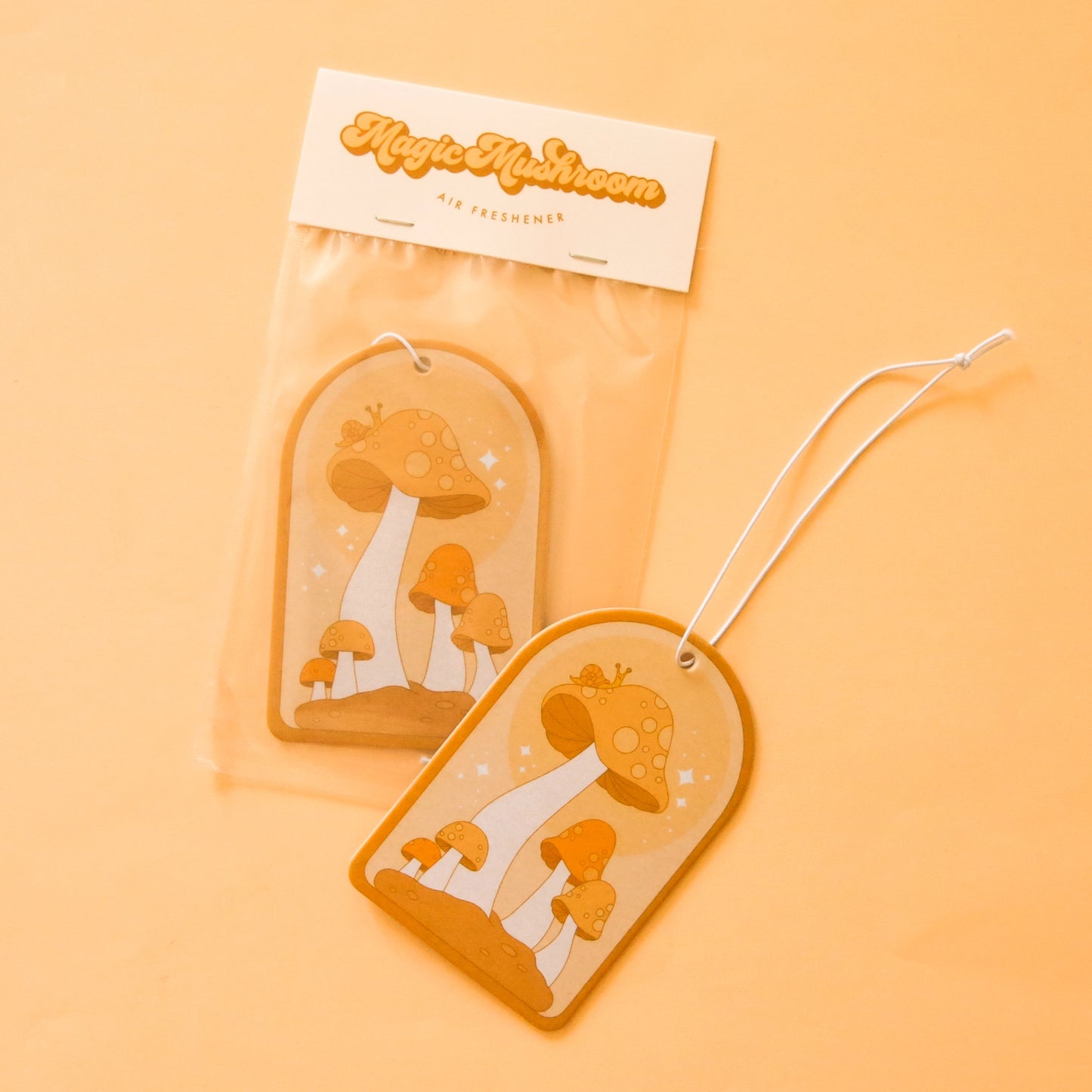 On a light orange background is an arched shaped air freshener with an orange mushroom design and a white elastic loop for hanging.