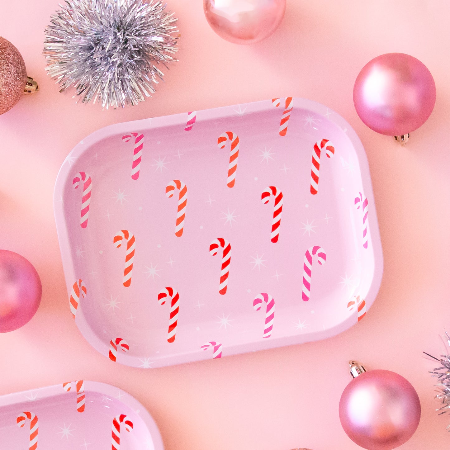 An aerial photo of a metal tray sits in the middle of page. The tray has pink, orange and red candy canes on it with white sparkle detail. The background of the image is a soft pink color. There are pink ornaments and tinsel ball decorations scattered around the tray.