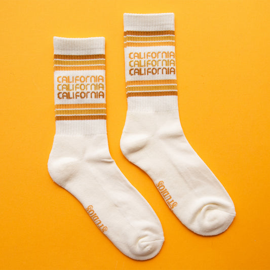 On a peach background is a white socks with rust, yellow and orange stripes and "california" printed three times on the ankle.'