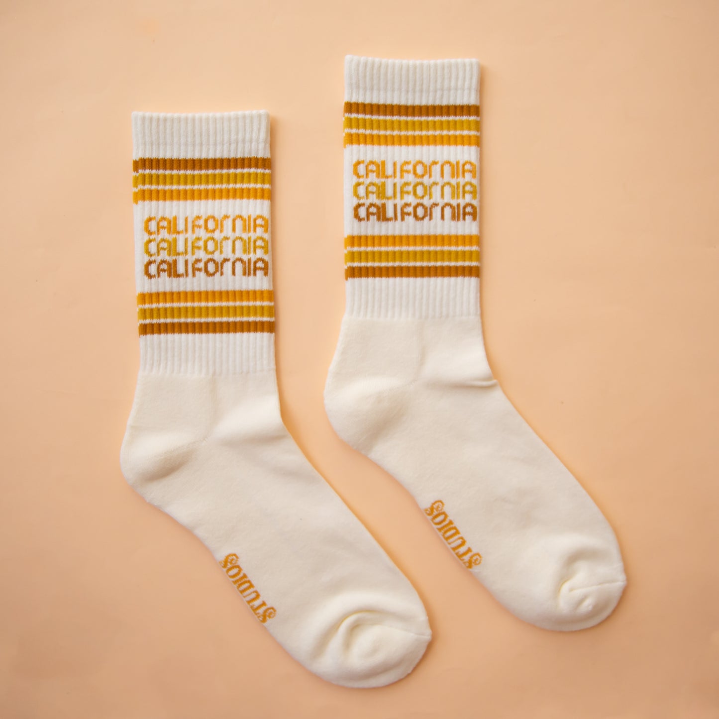 On a peach background is a white socks with rust, yellow and orange stripes and "california" printed three times on the ankle.