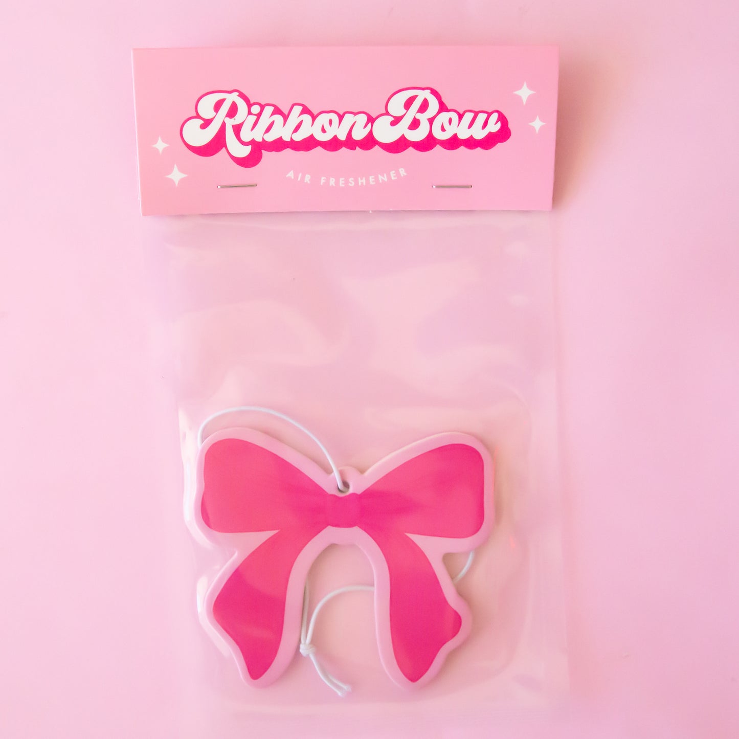 A hot pink bow with light pink trim air freshener is shown against a light pink to peach gradient background.