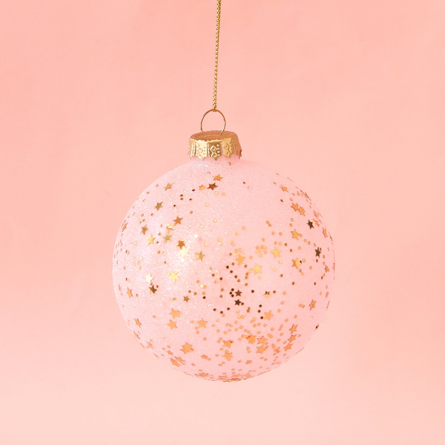 On a peachy background is a light pink ball ornament with small gold stars all over.