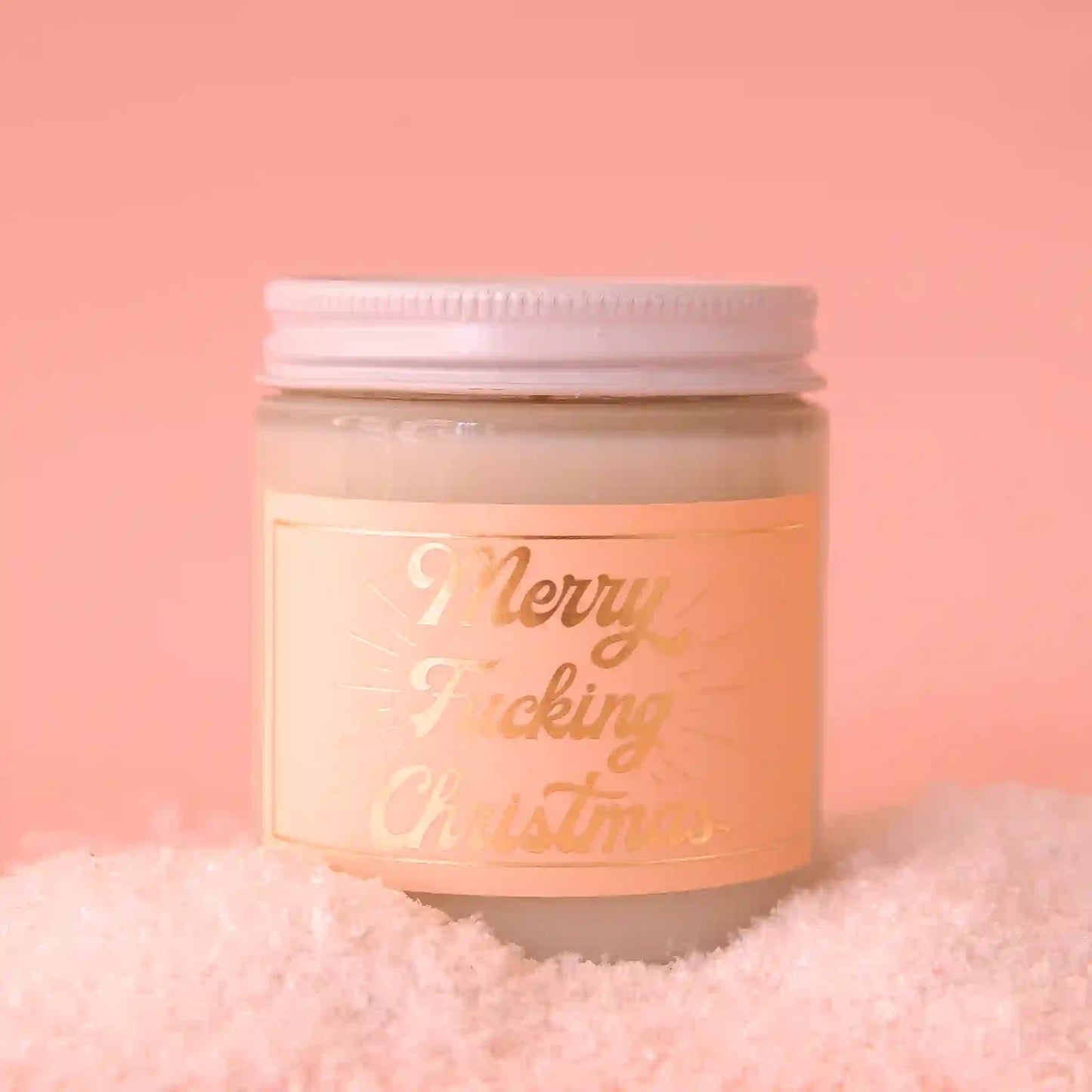 On a pink background is a clear glass jarred candle with a white lid and a peachy label that has gold text reading, "Merry Fucking Christmas".