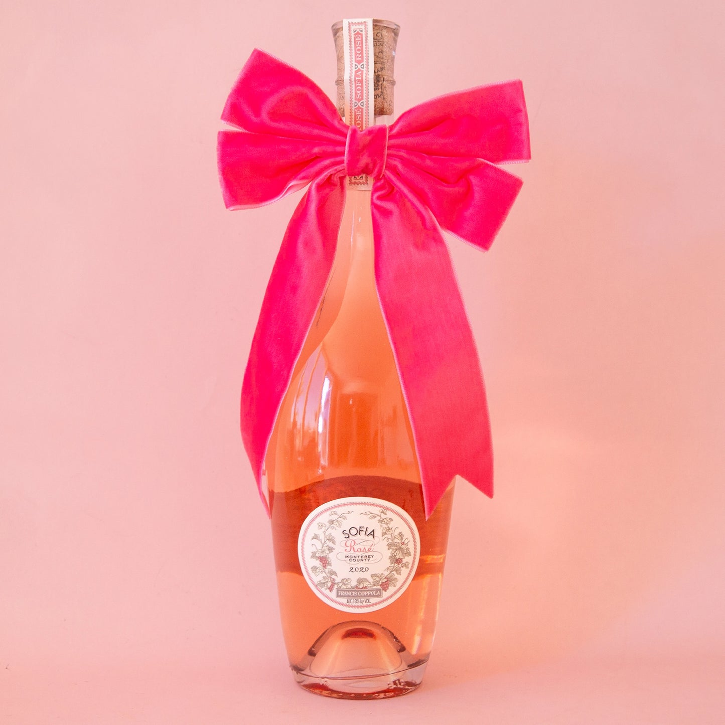 On a peachy background is a wine bottle with a large velvet pink bow.