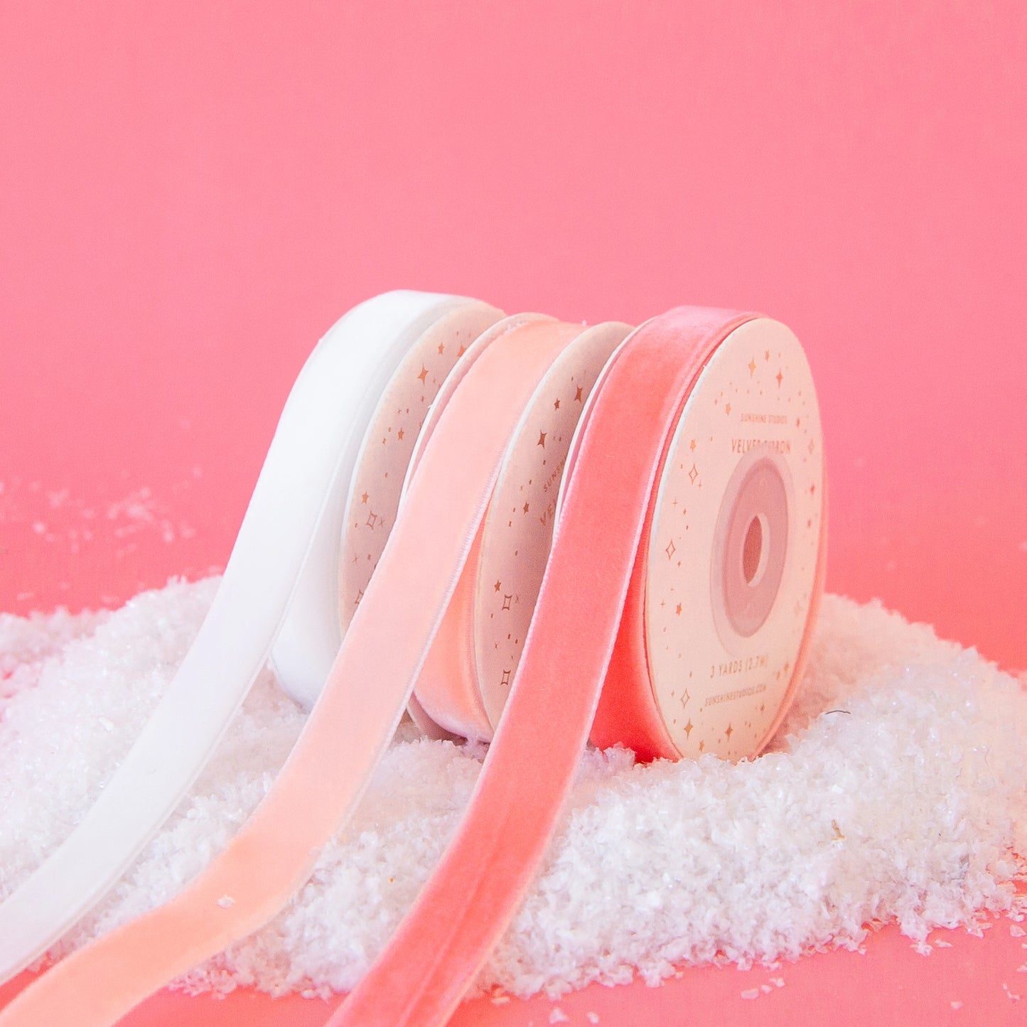 On a pink background is three spools of velvet ribbon in three different shades ranging from white to a peachy pink.