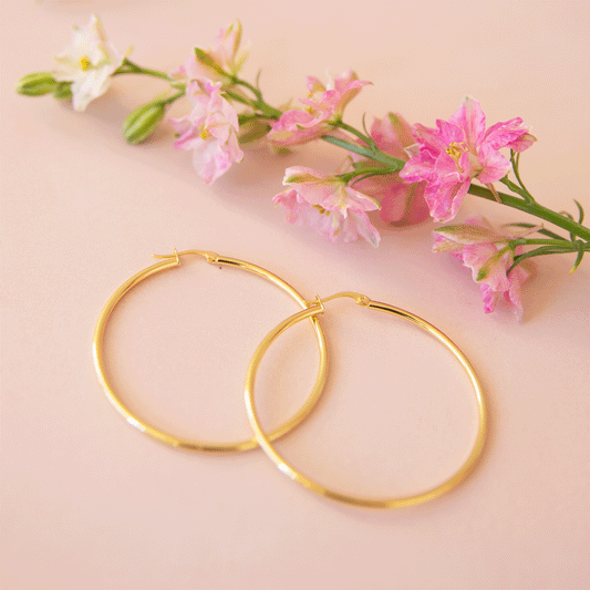 On a tan background is a pair of thin gold hoop earrings next to a stem of pink flowers.