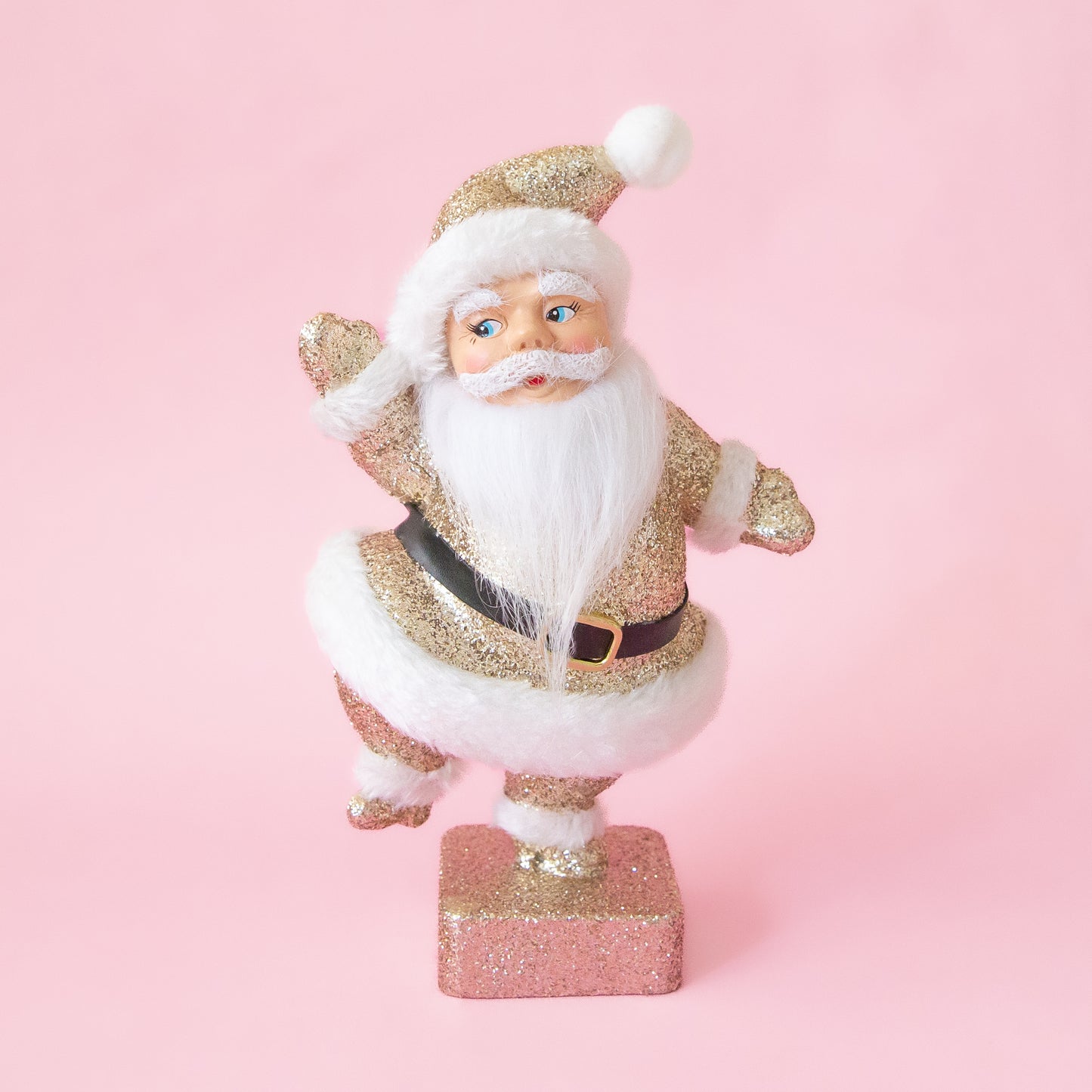 A vintage inspired Santa statue covered in gold glitter. He wears a white beard and white fur details. The photo is on a pink background.