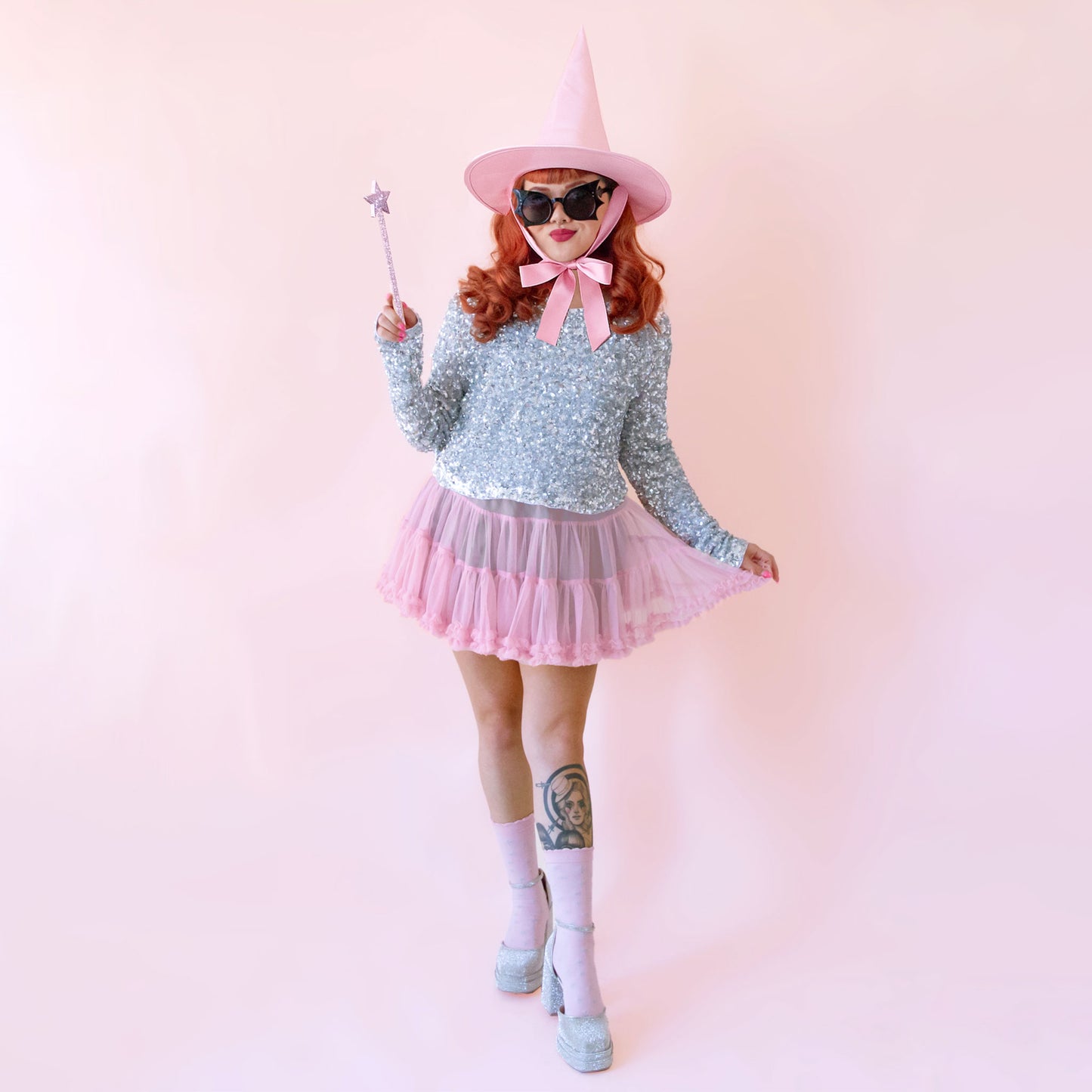 A model dressed in a pink skirt and silver sequin top stands in the middle of frame holding a star shaped wand and wearing a pink witch hat.