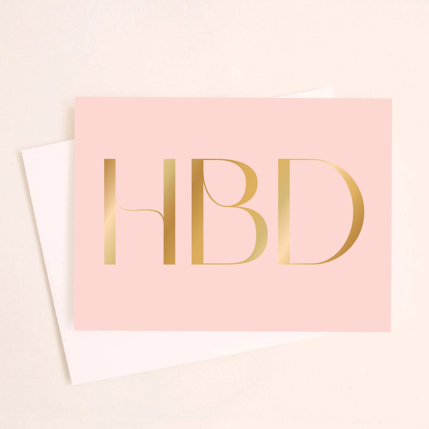 On an ivory background is a light pink greeting card with large gold foil text in the center that reads, "HBD" in all caps. Also included is a white envelope.