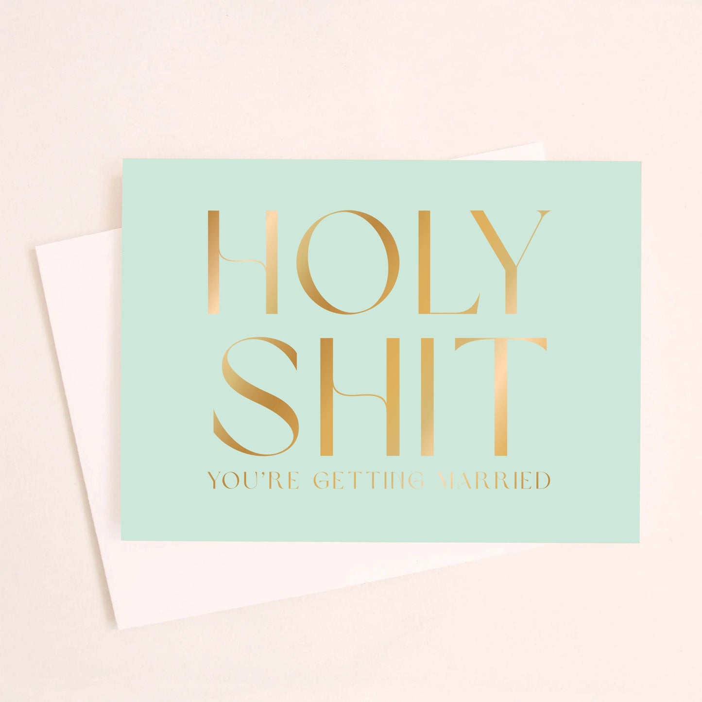 On an ivory background is a teal greeting card that reads, "Holy Shit You're Getting Married" in gold foil letters and a matching envelope beside it.