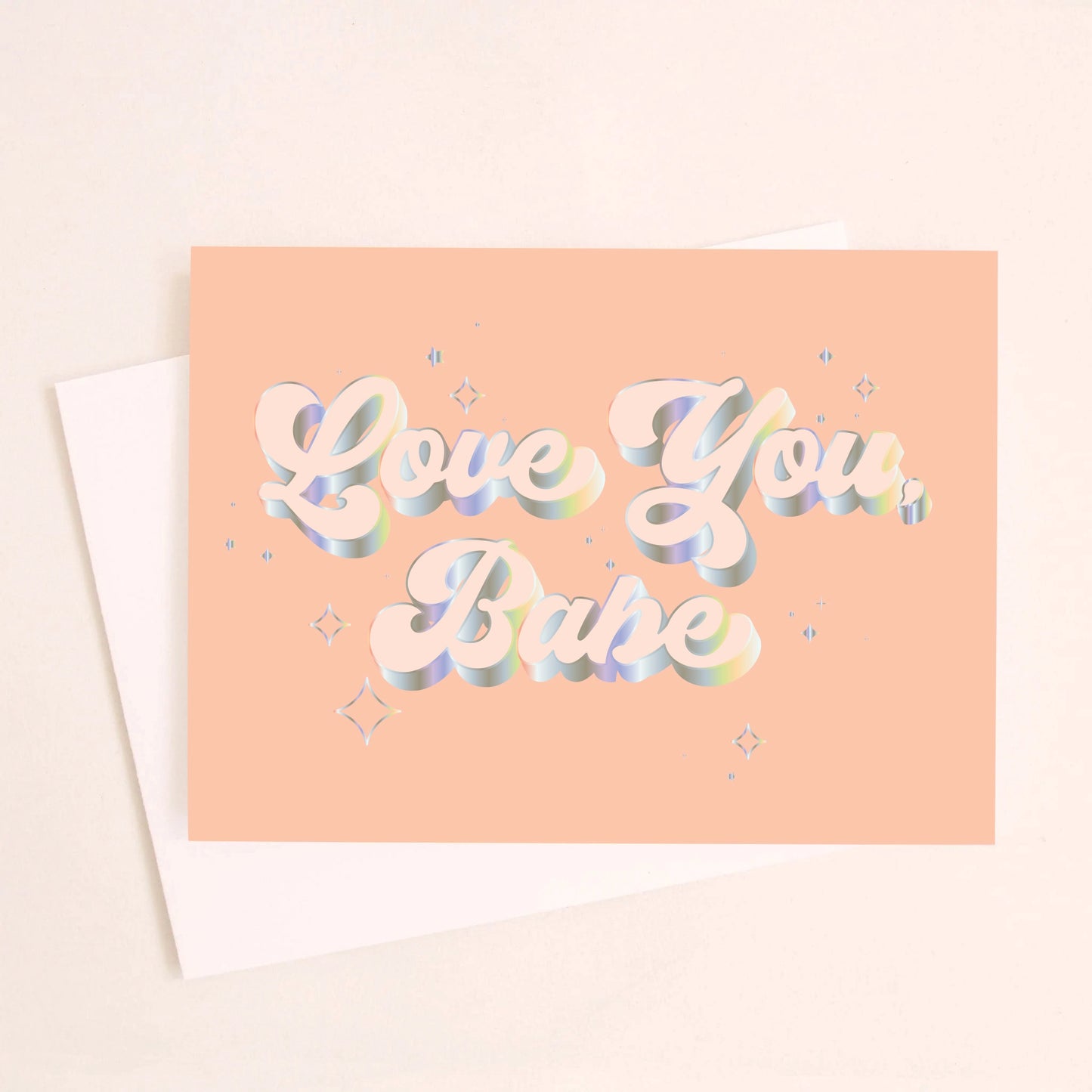 On a light peach background is a peachy greeting card with "Love You Babe" text in the center that is outlined with holographic foil detailing. Also included is a white envelope.