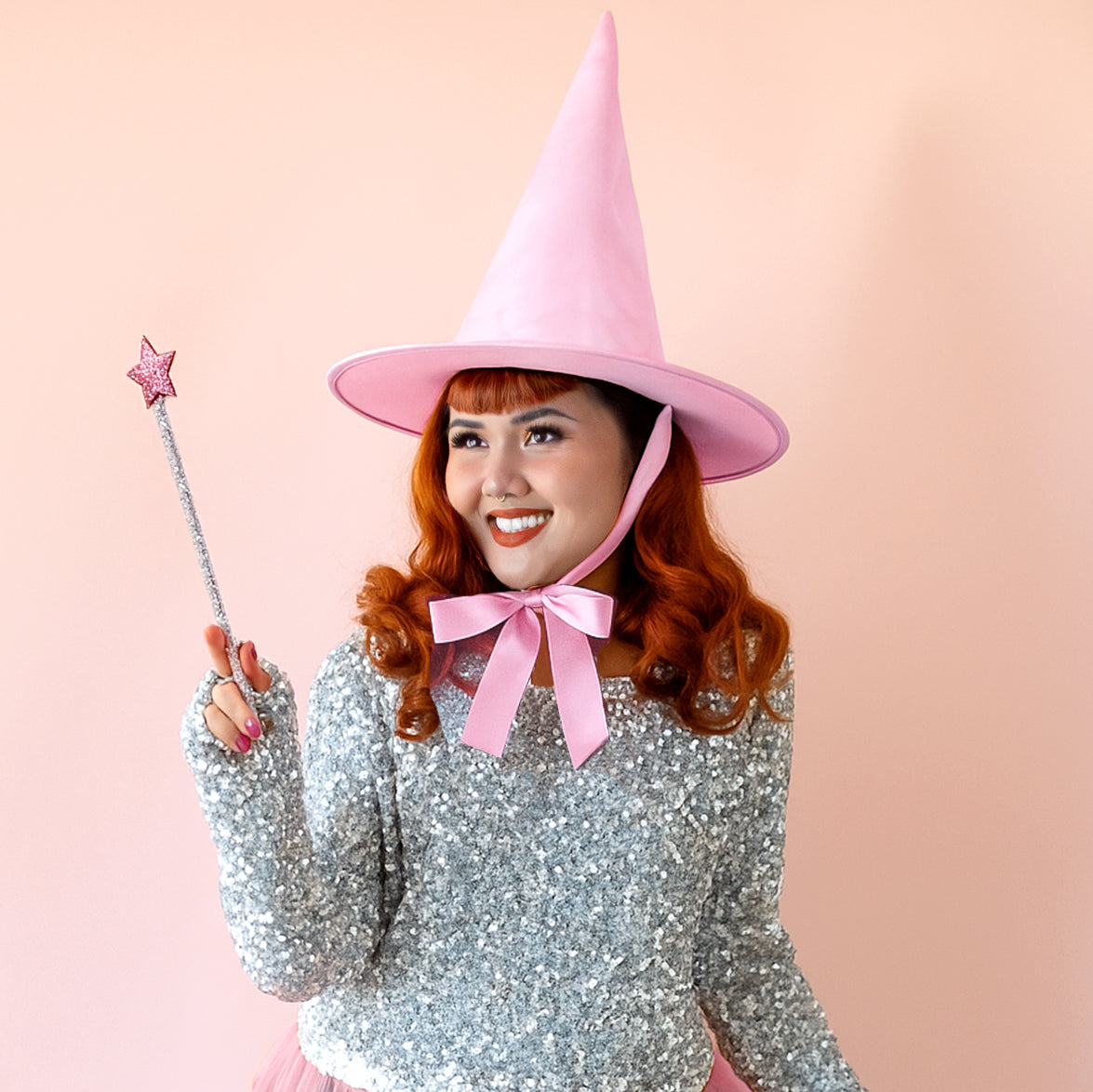 Red headed model wears a pink witch hat with a bow and holds a wand.