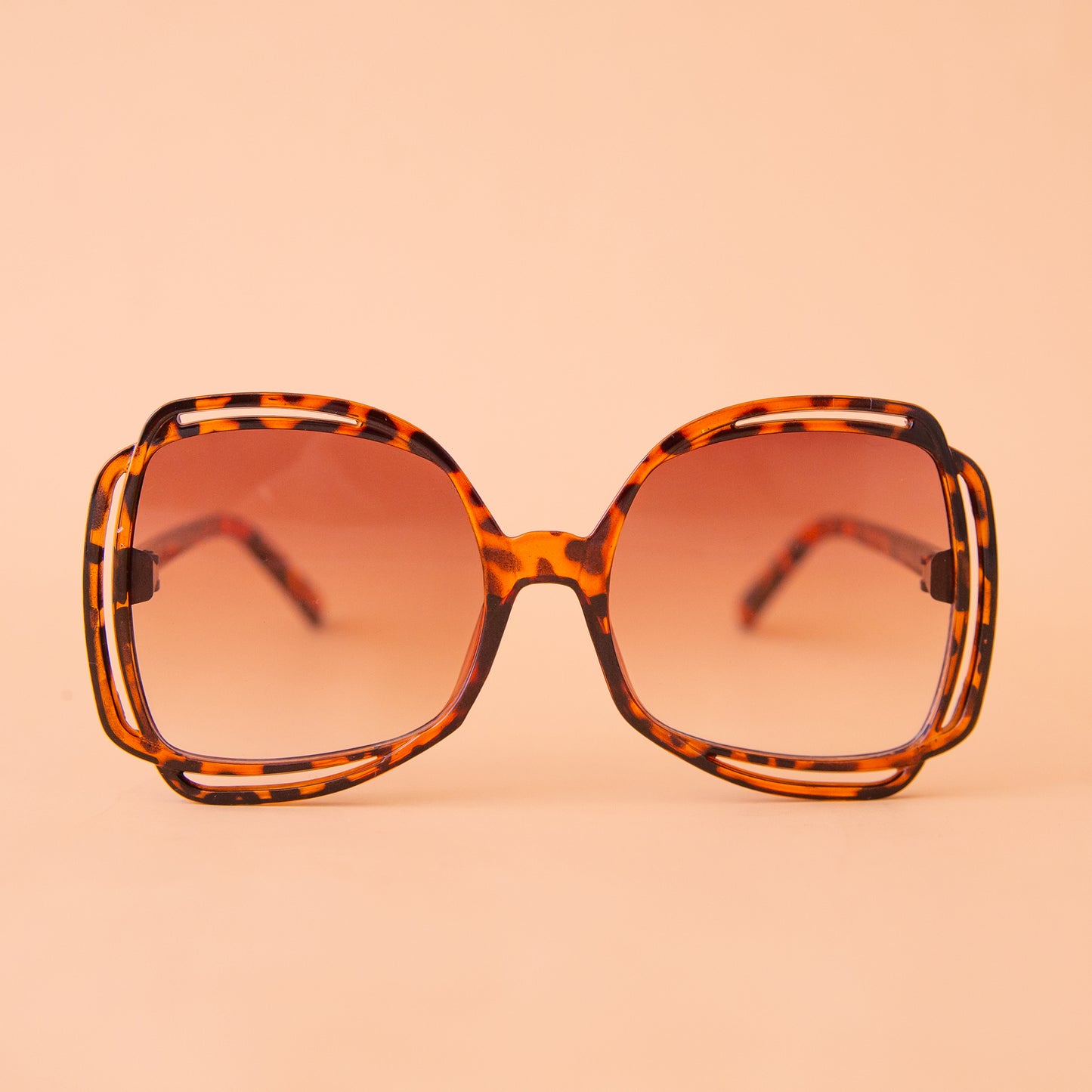 On a peachy background is a pair of tortoise sunglasses with a round frame and a brownish pink lens.