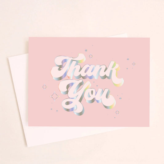 On an ivory background is a light pink card that reads, "Thank You" with in white text with holographic outlining. 