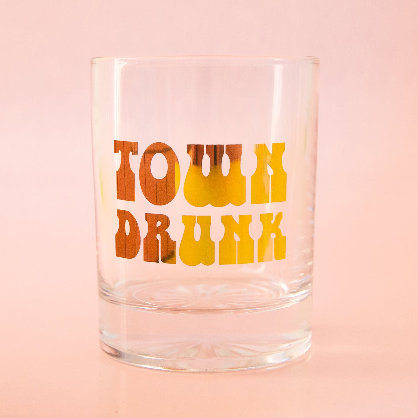 Glass tumbler with gold foil writing, spelling"town drunk" is shown against a light pink background
