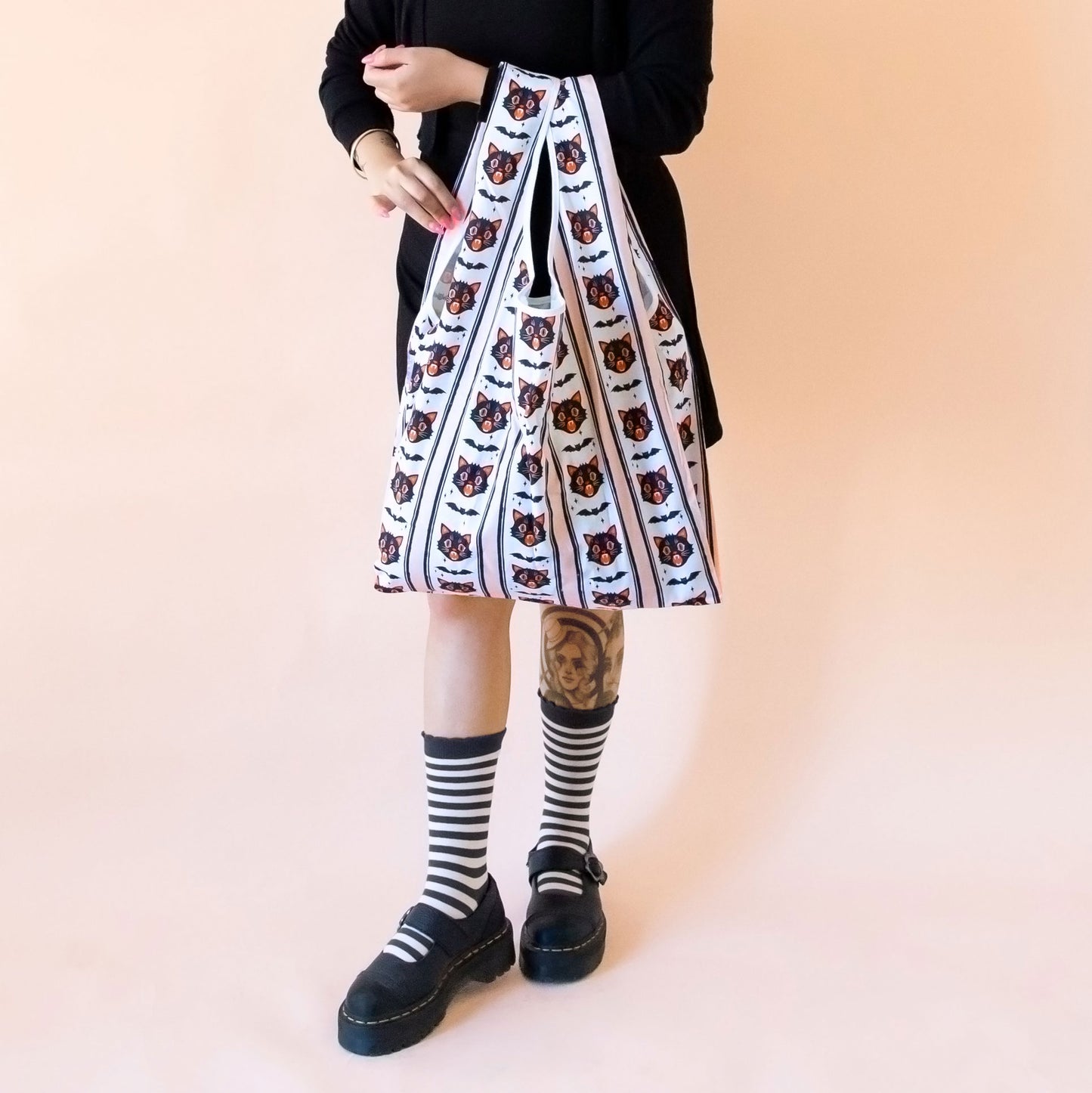 Person standing in black and white striped socks holding a bag that has vintage black cat faces and bats designed on it. The figure is wearing black clothing.
