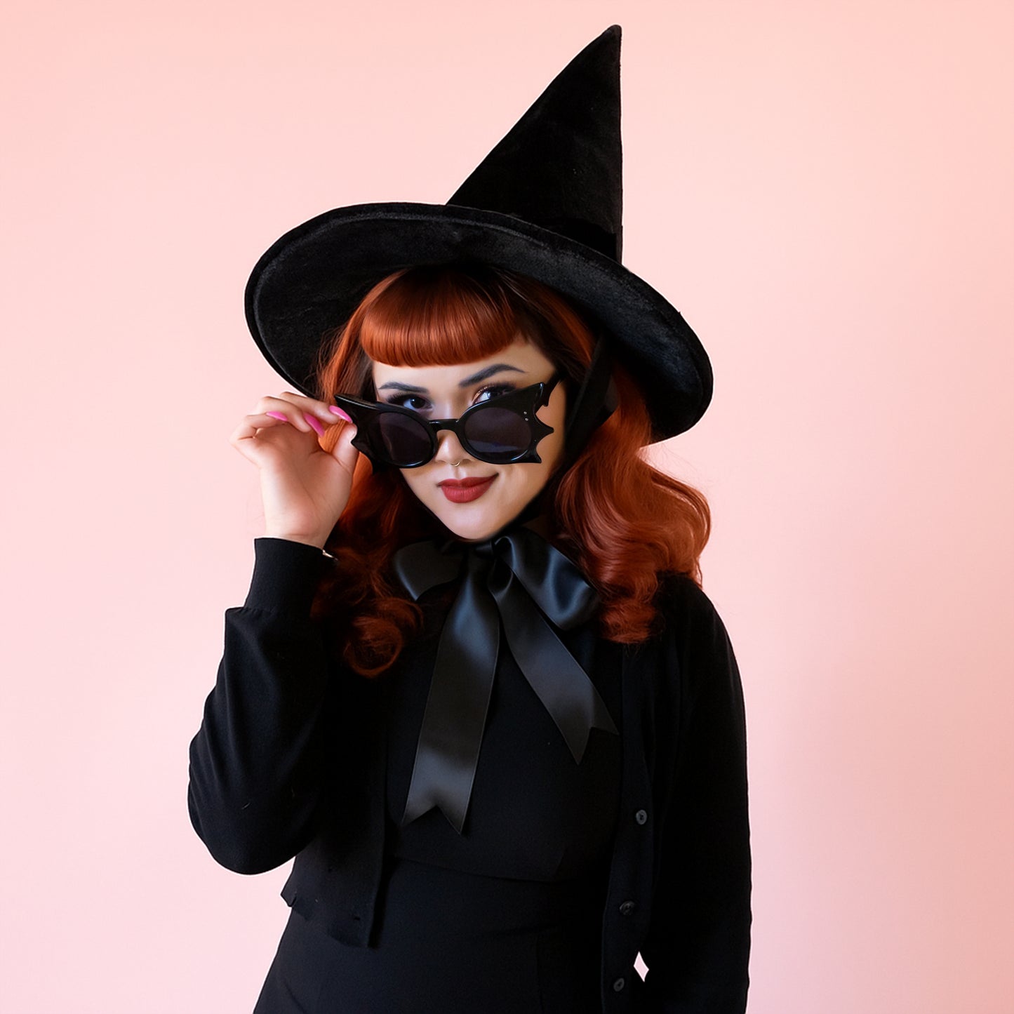 A red headed woman wears black bat shaped sunglasses and a tall black witch hat with a bow at the neck. She is wearing an all black outfit.