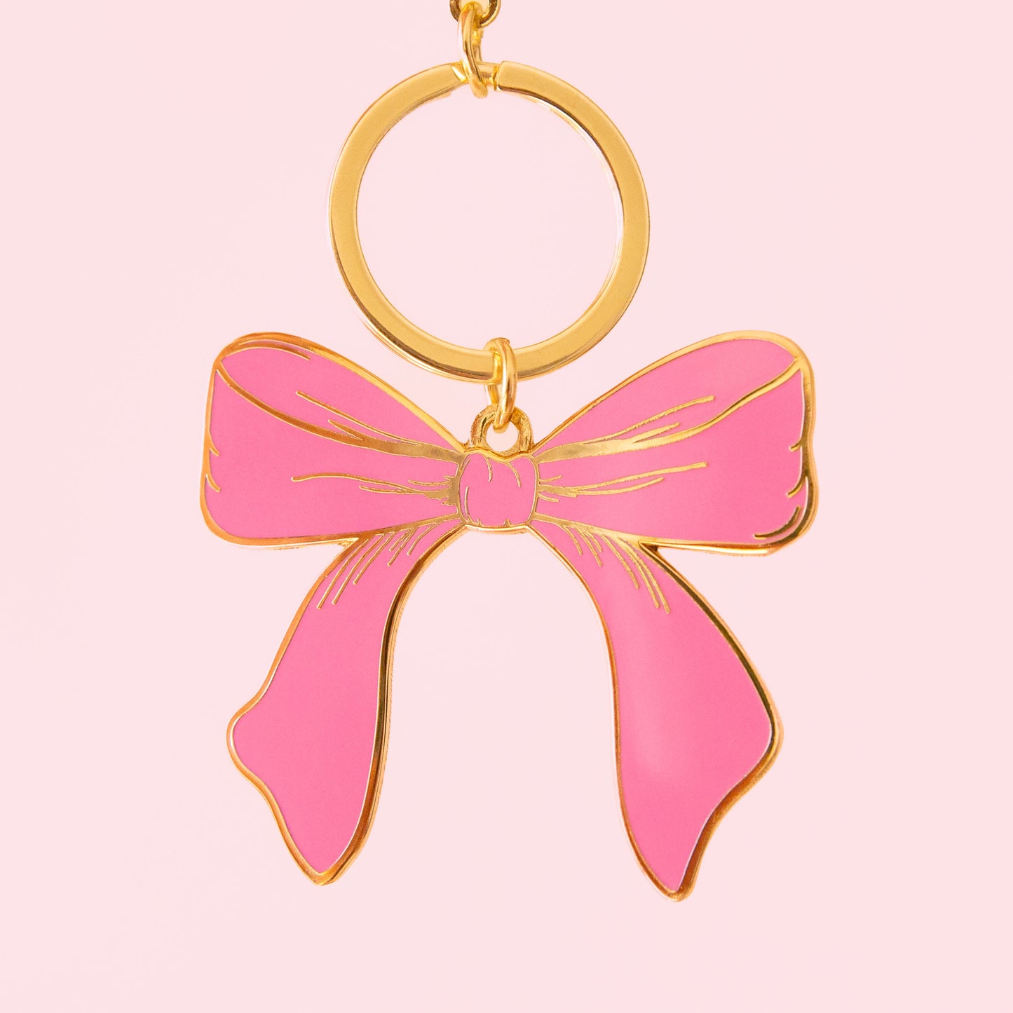 A pink enamel bow keychain with gold hardware hangs on a soft pink background.