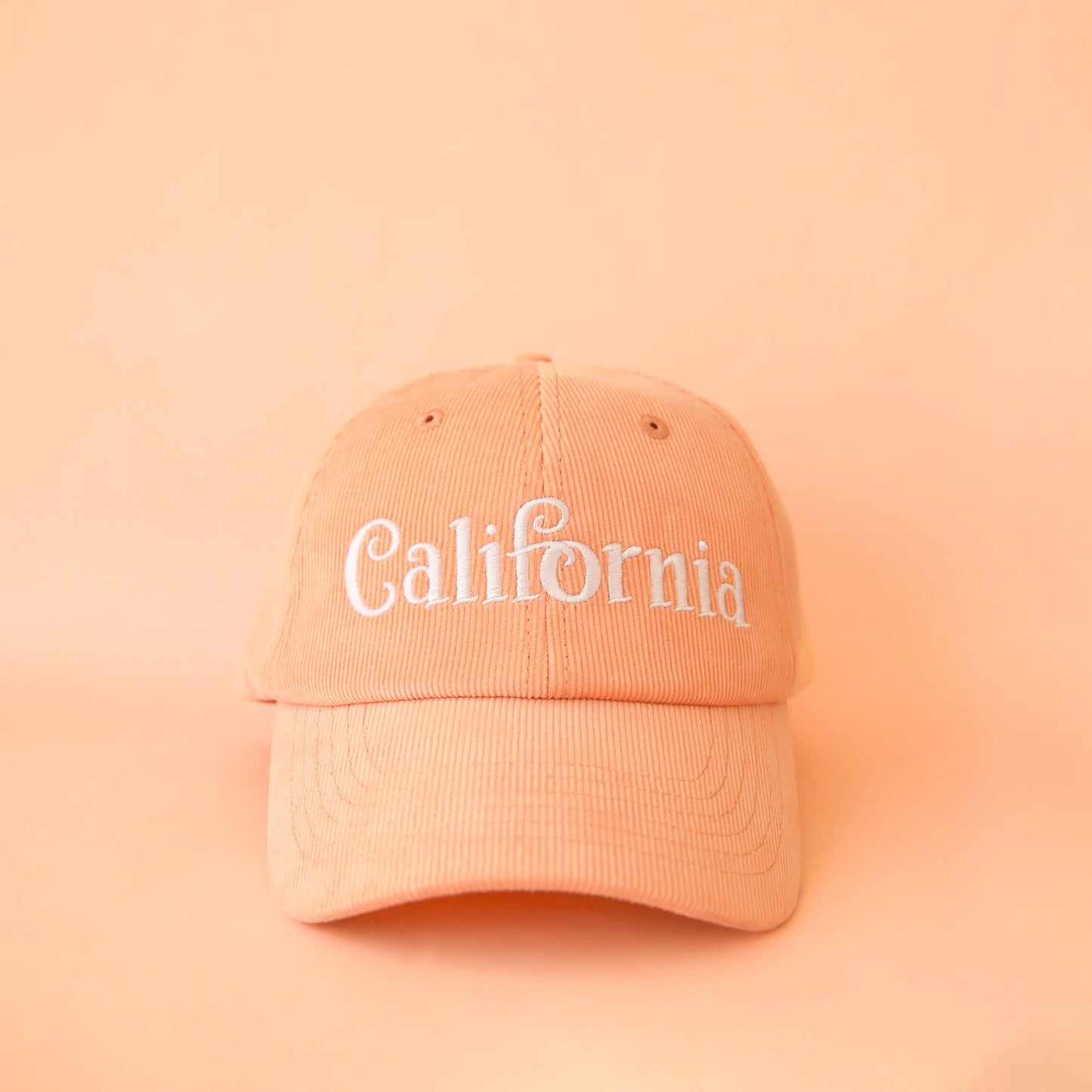 On a peach background is a peach colored baseball hat with a corduroy material and white embroidered text across the front that reads, "California".