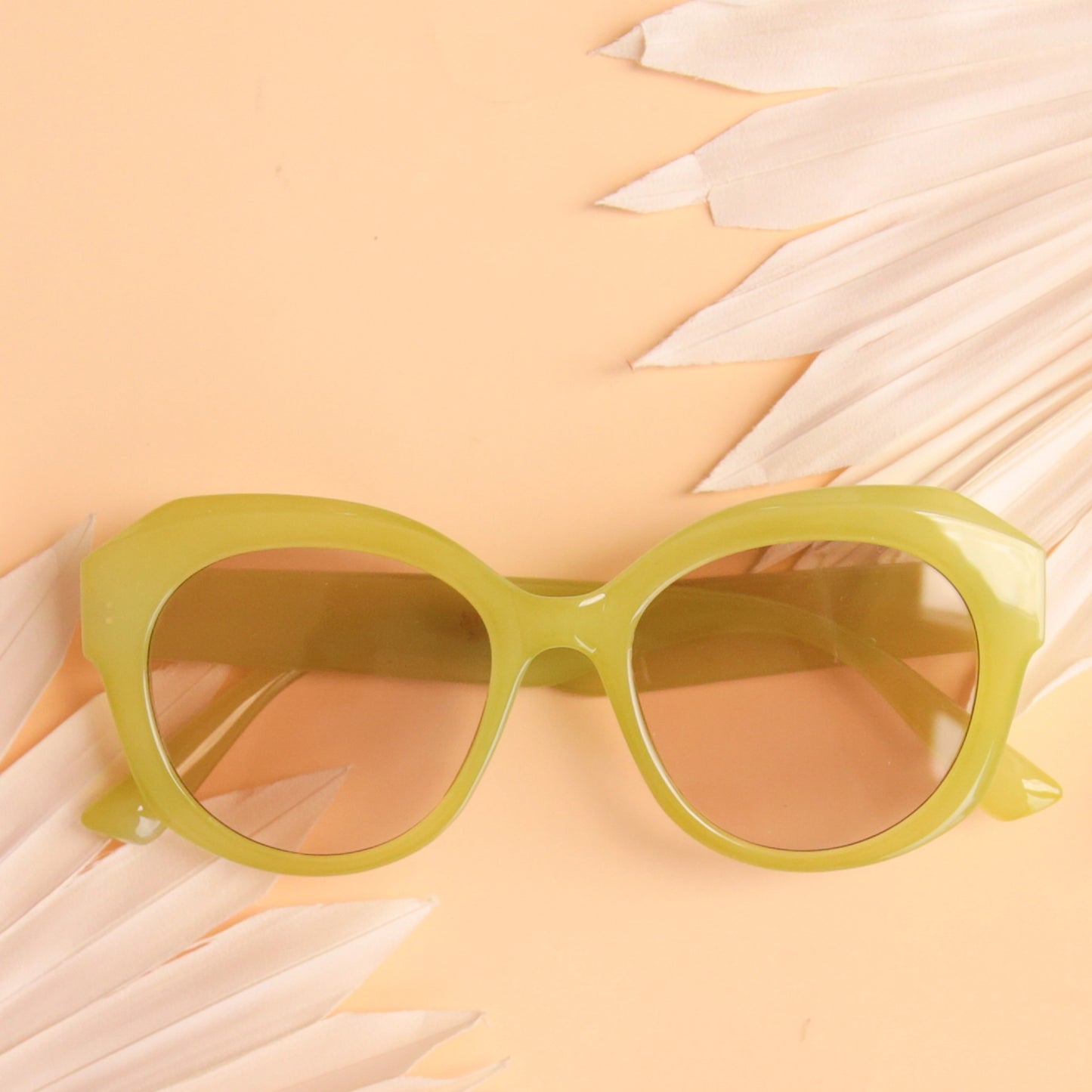 On a peachy background is a round pair of light green sunglasses with a light brown lens.