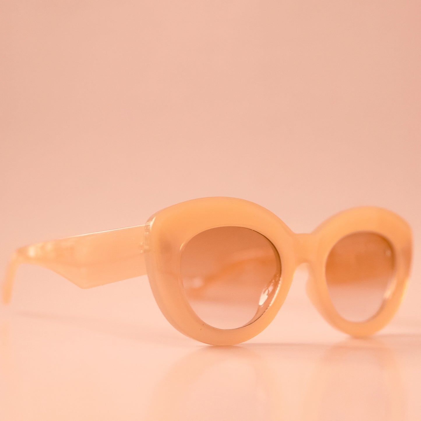 On a peachy background is the Gemma Sunglasses in the shade Amber. They have a rounded shape with a slight cateye flair at the corners. The frame material is a durable amber / peachy colored plastic and the lenses are a similar tone.