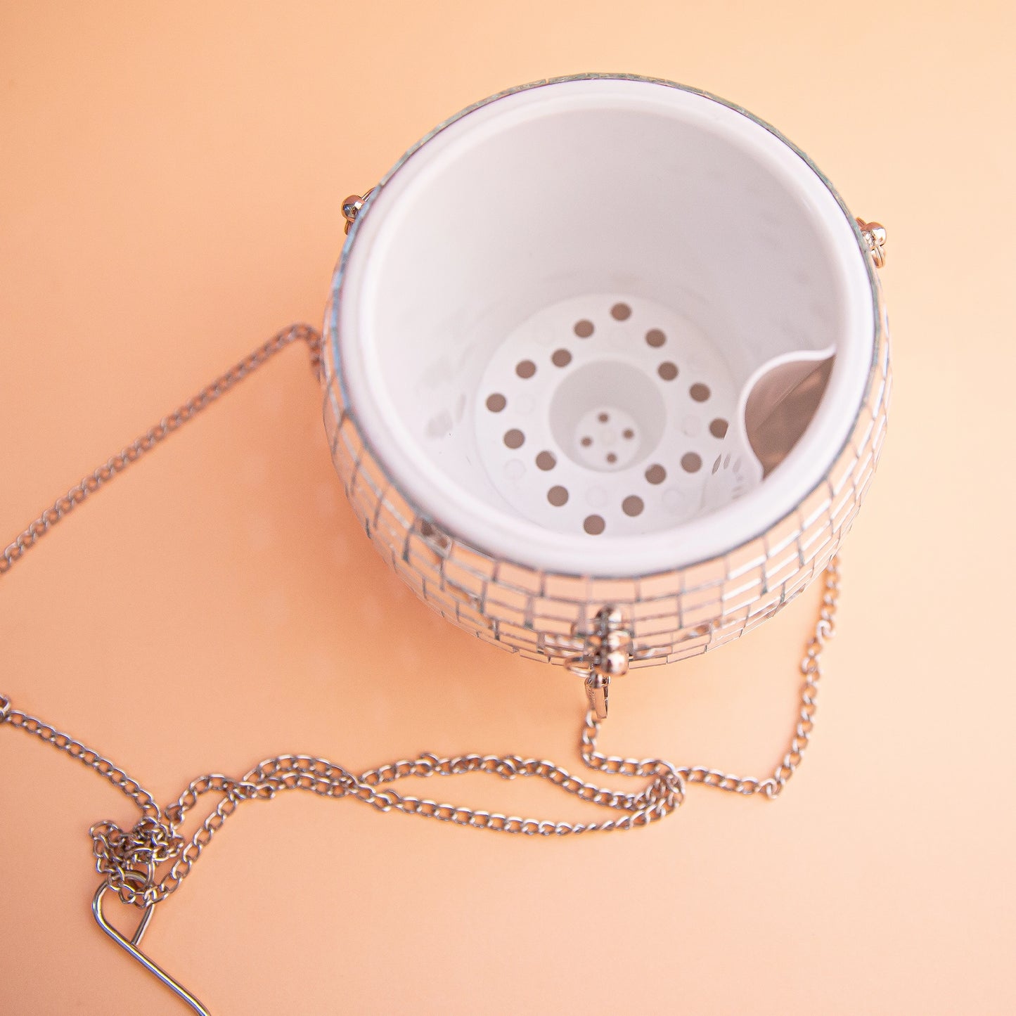 On a peachy background is a silver mirrored glass disco ball shaped planter with a silver chain hanger with a drainage system inside.