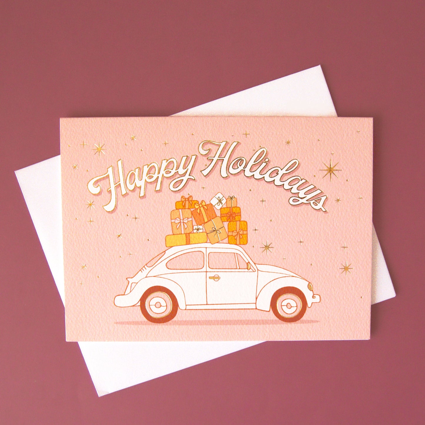 On a pink background is a card with a VW bug with gifts on top and text arched above it that reads, "Happy Holidays".