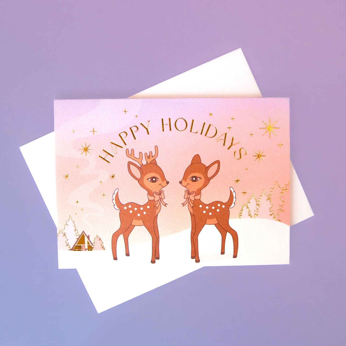 On a purple background is a greeting card with two brown retro deer graphics along with gold foiled details and text that read, "Happy Holidays".