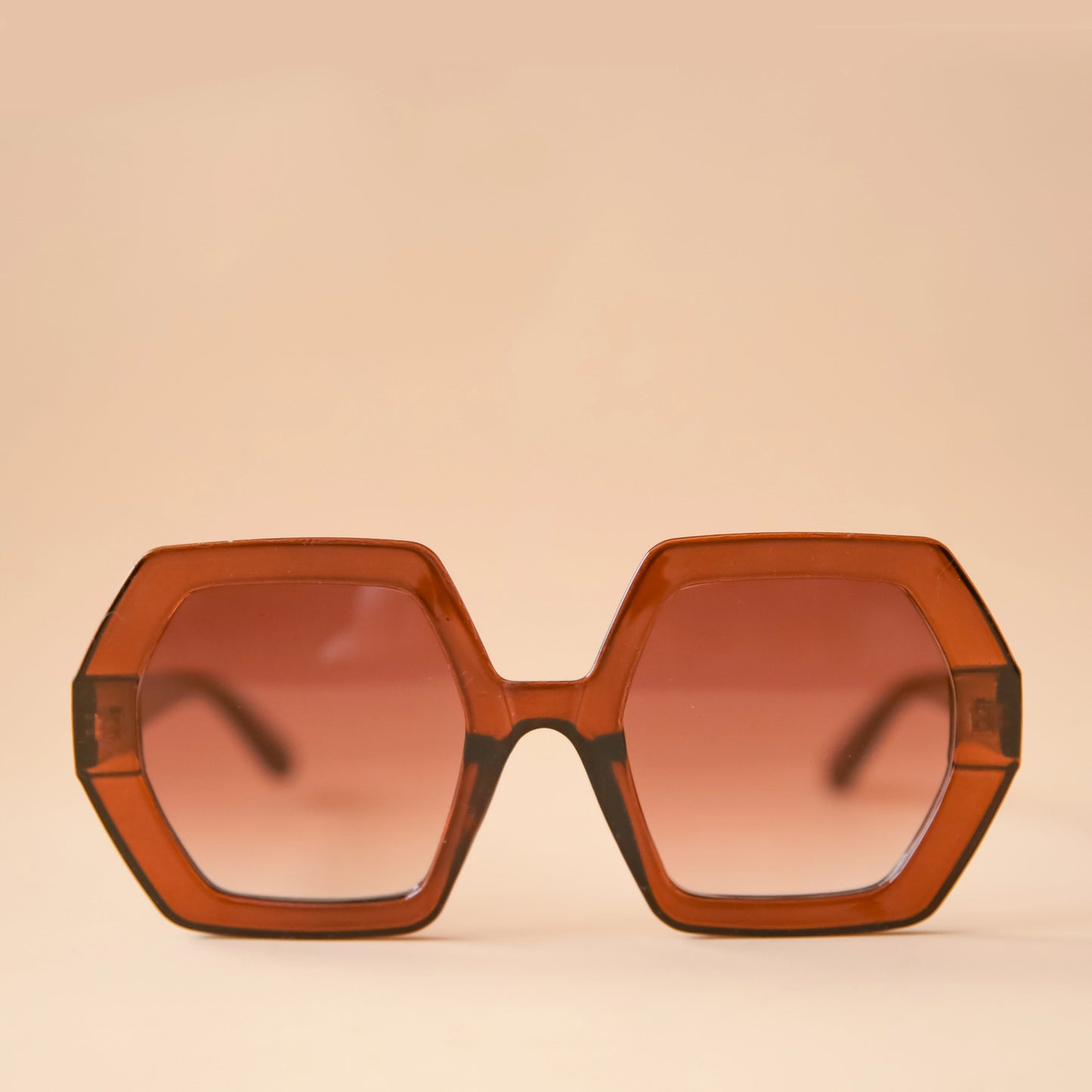 On a peachy background is a pair of hexagon shaped sunglasses in a cognac brown color.