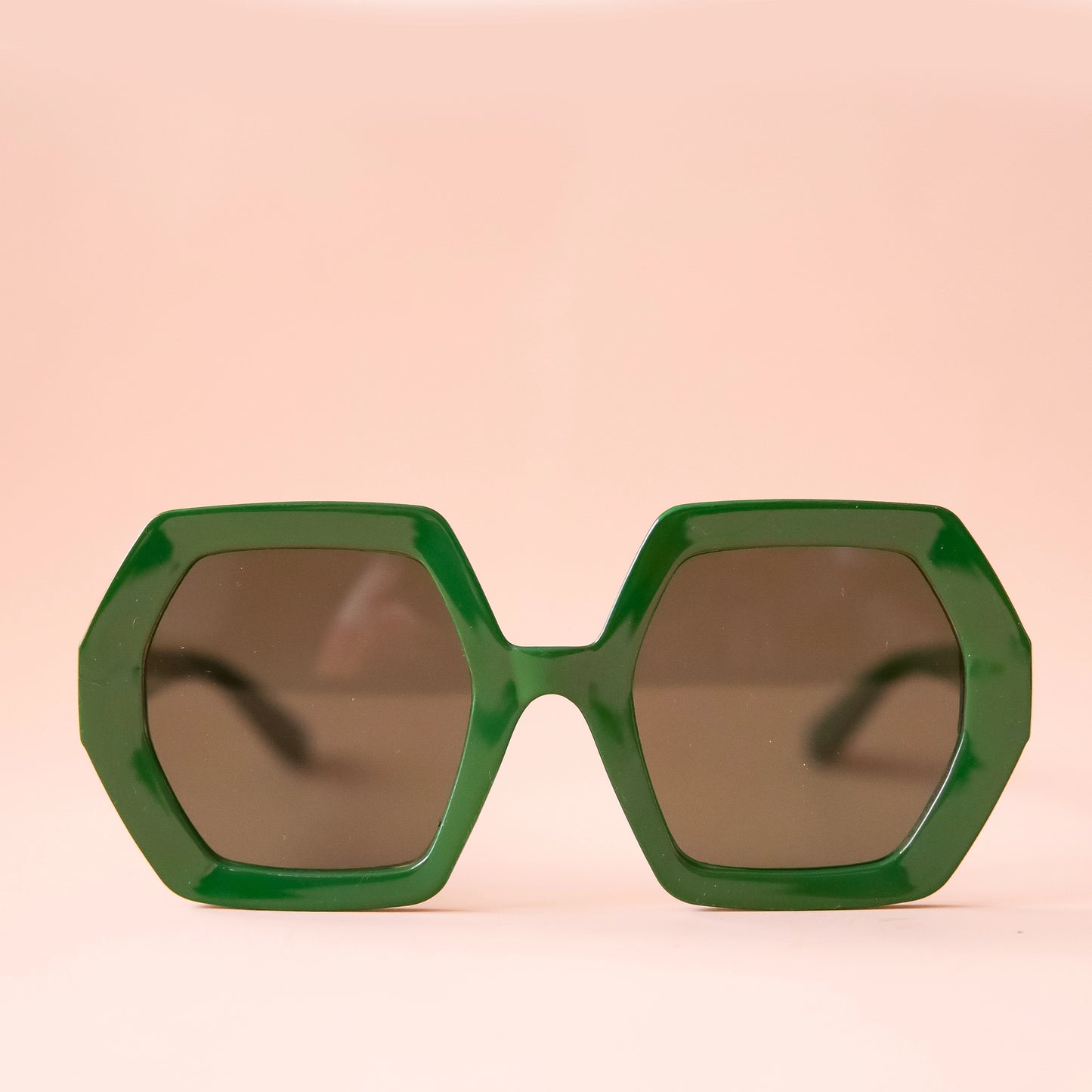 On a peachy background is a pair of hexagon shaped plastic sunglasses in an emerald green color with a dark brown lens.