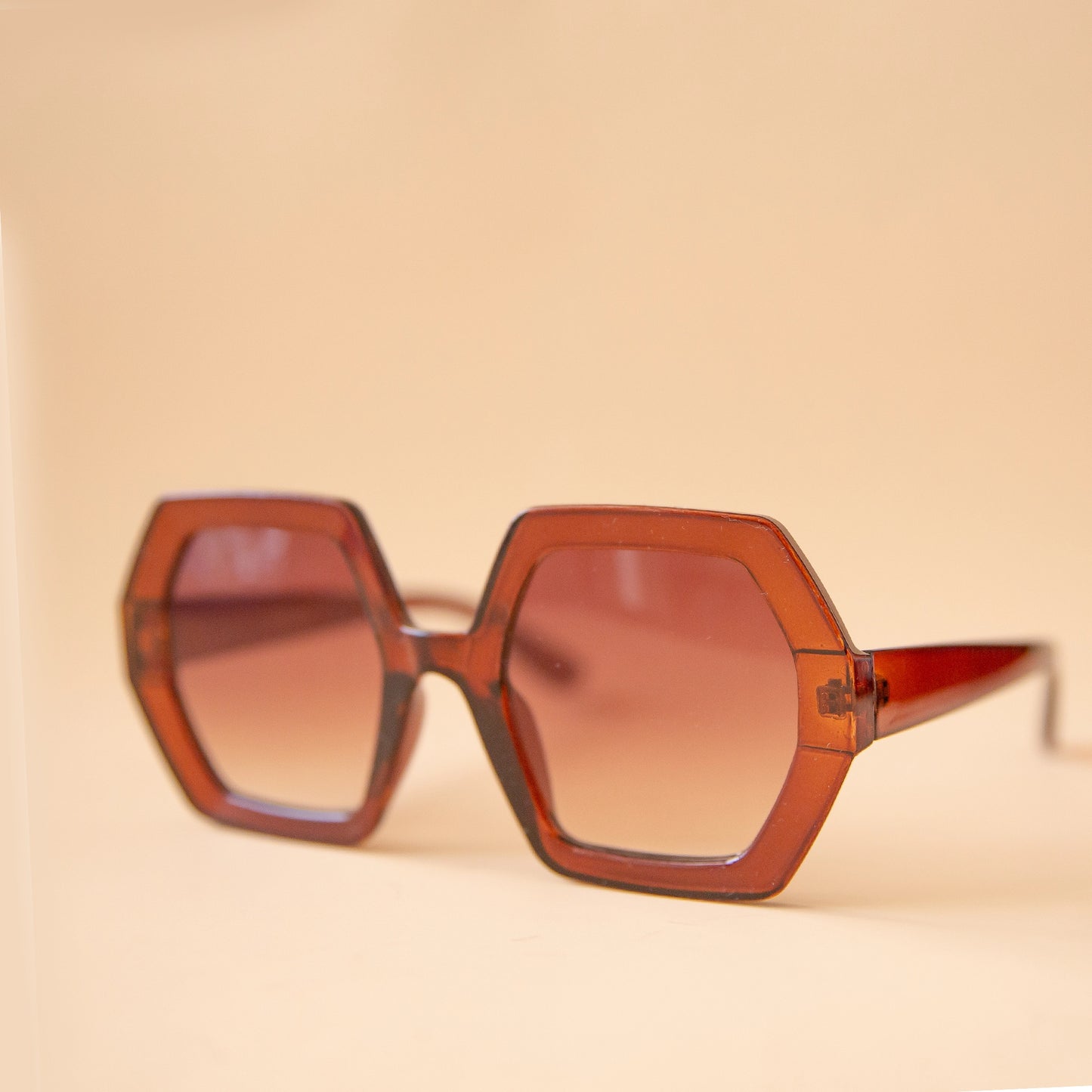 On a peachy background is a pair of hexagon shaped sunglasses in a cognac brown color.