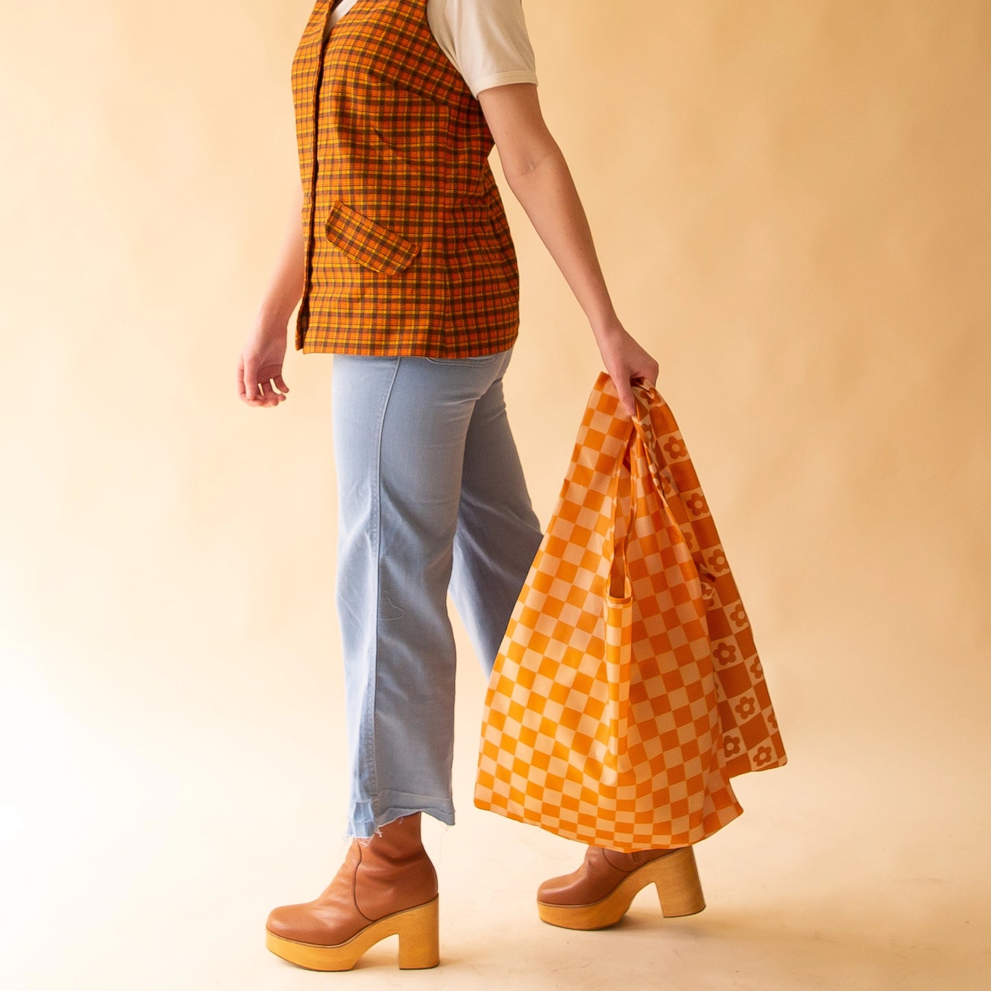 On a peach background is a model holding two nylon reusable bags, one being the playa check which is a dark orange checkered print.
