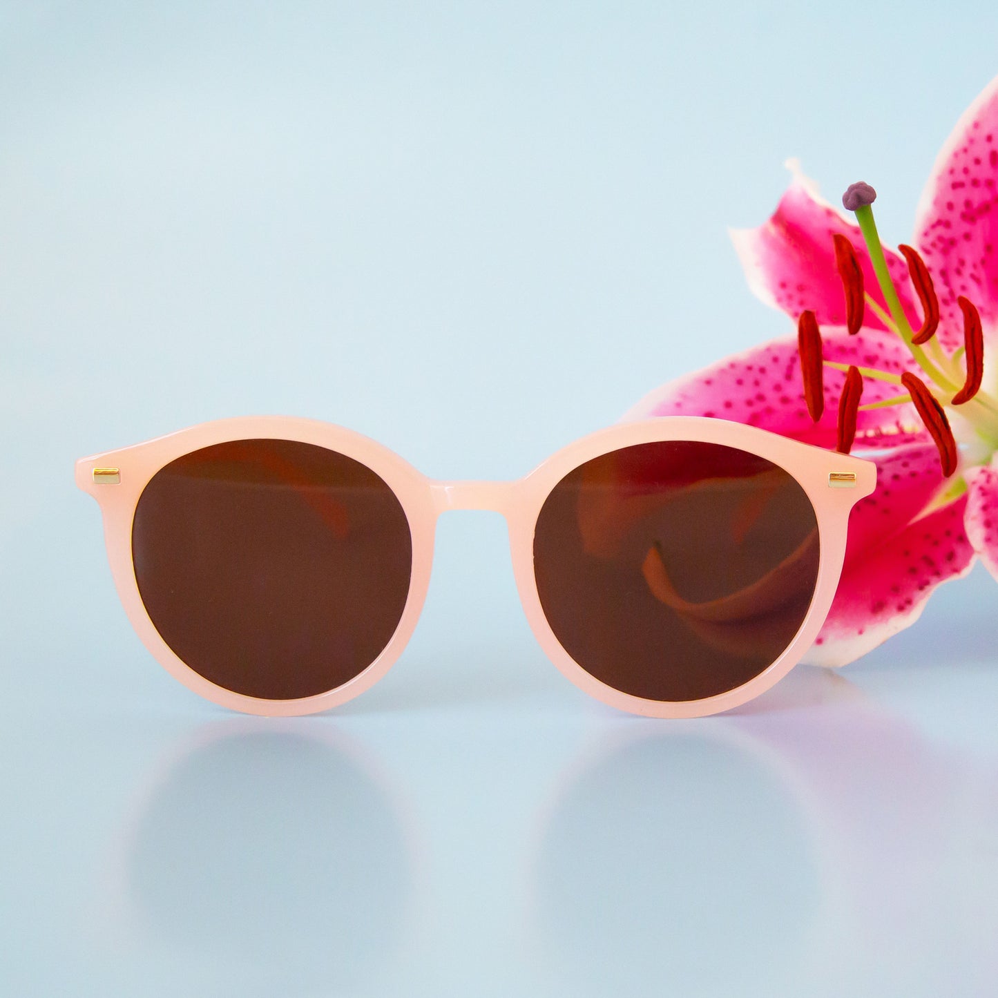 On a pink backgrounds a pink framed pair of round sunglasses with brown lenses.