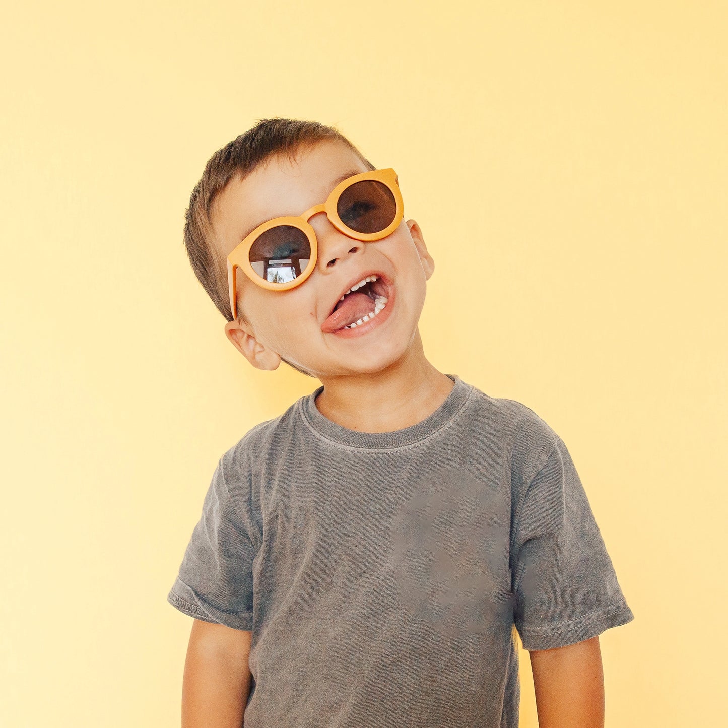 On a light yellow background is. children's model wearing a pair of round sunglasses in a yellow shade.