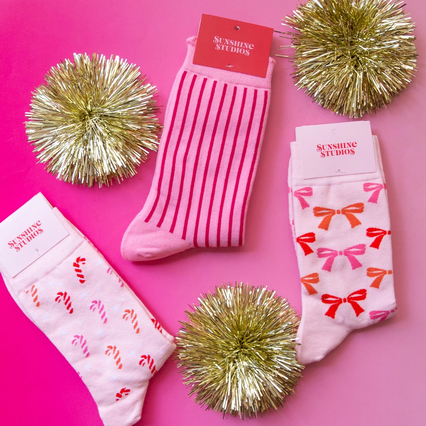 Three pair of pink socks with holiday details- bows, stripes and candy canes and three gold sparkly metallic decorative puffs on a pink background.