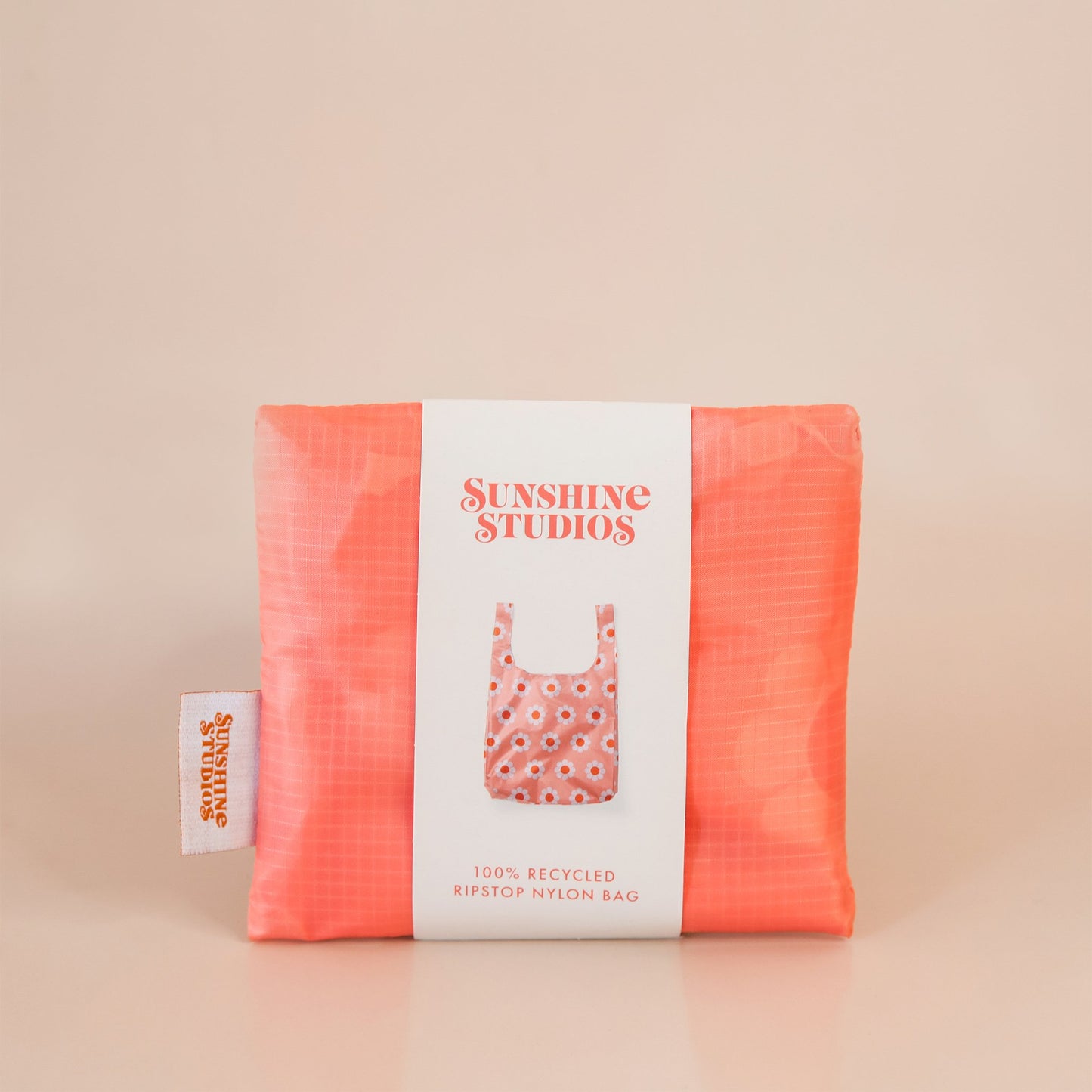 Peach reusable bag folded tightly into a square. The bag is wrapped in a white band that reads ’sunshine studios'. Under the text is a picture of the reusable bag. On the left side of the bag is a white tag with bright orange text that reads ’sunshine studios.'