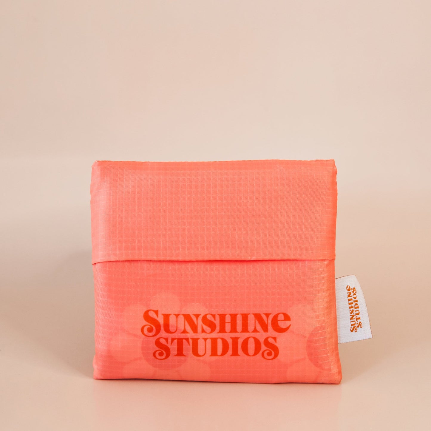 In front of a tan background is a square peach pouch. At the bottom is orange text that reads ’sunshine studios.’ On the right side is a white tag with orange text that reads ’sunshine studios.’