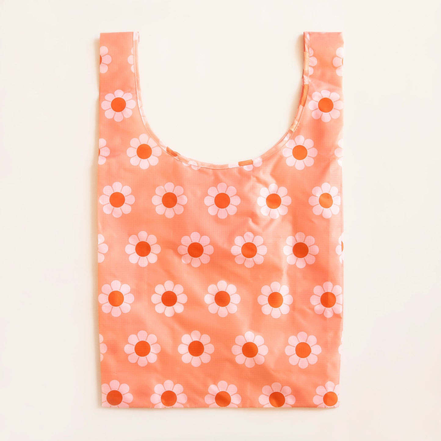 Peach reusable bag covered in a print of simple flowers with white petals and orange centers. The bag is positioned flat on a table and has a 'U' shape between two handles.
