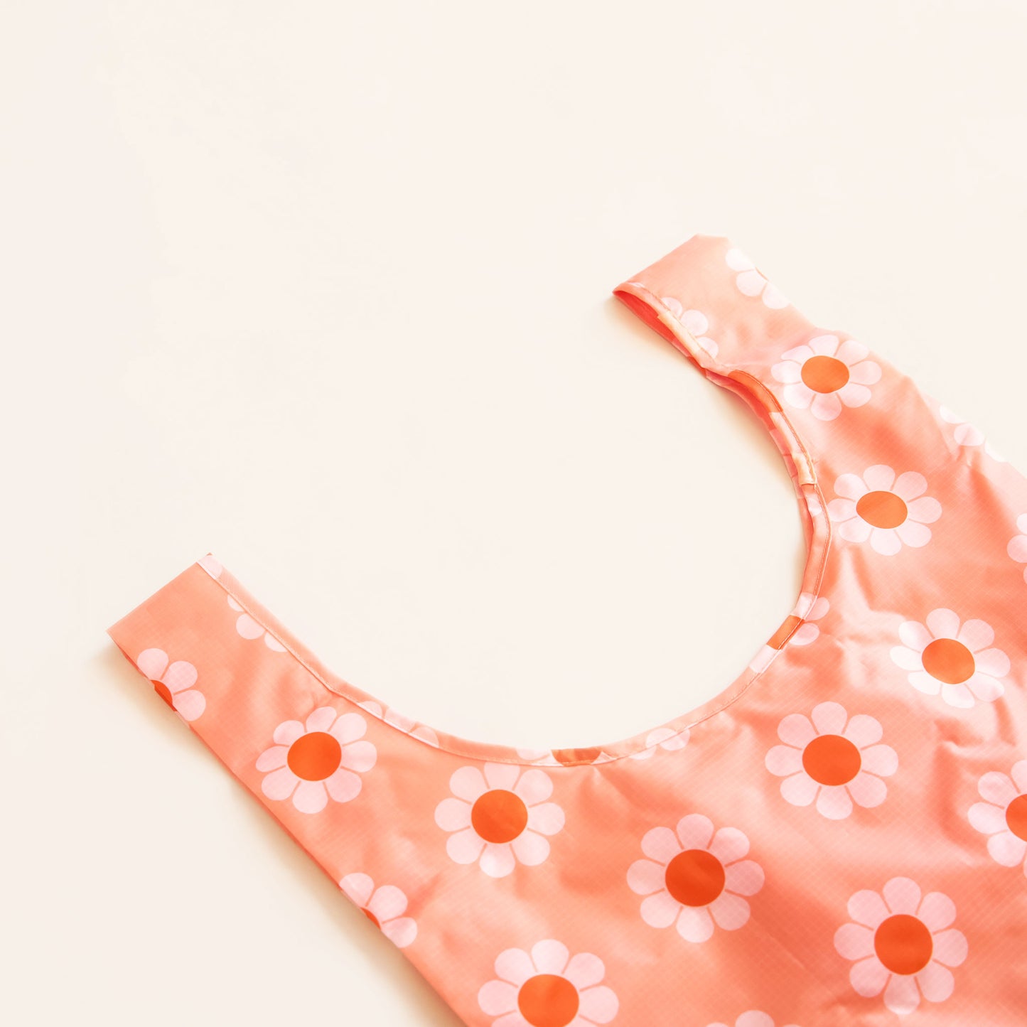 Zoomed in view of peach reusable bag covered in a print of simple flowers with white petals and orange centers. The bag is positioned flat on a table and has a 'U' shape between two handles.