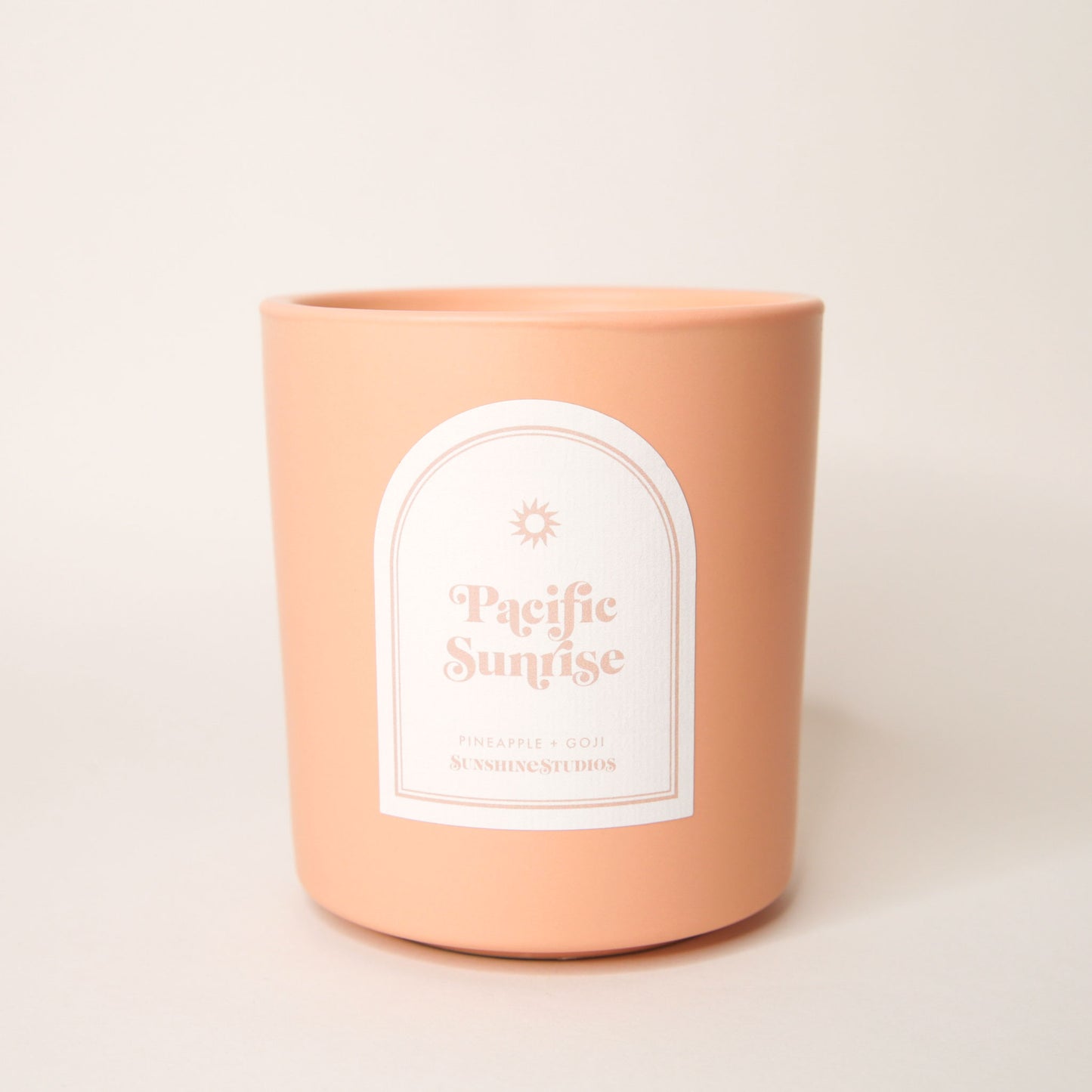 Pastel orange candle with an arched label that reads 'Pacific Sunrise, pineapple + goji, sunshine studios' in soft pink lettering. A small sun design sits above the text.