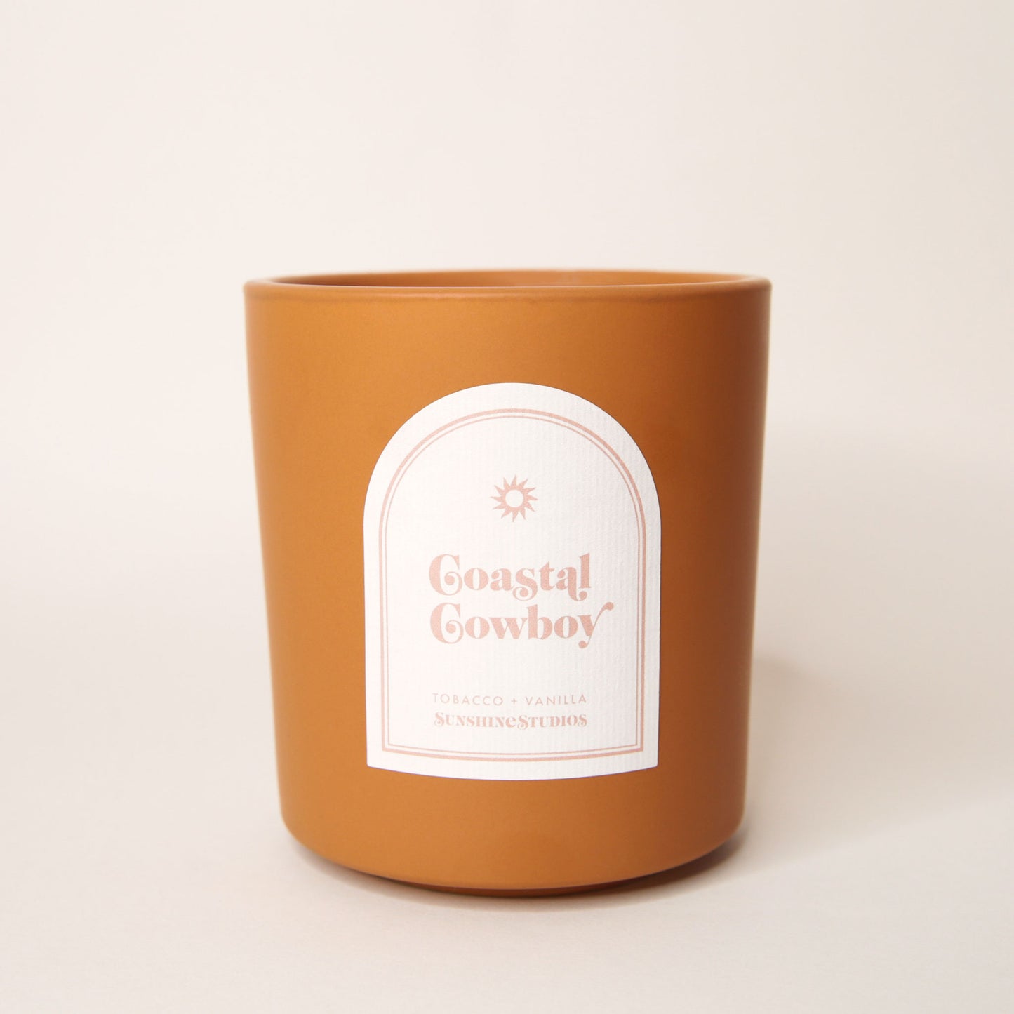 Pumpkin orange candle with an arched label that reads 'Coastal Cowboy, tobacco + vanilla, sunshine studios' in soft pink lettering. A small sun design sits above the text.